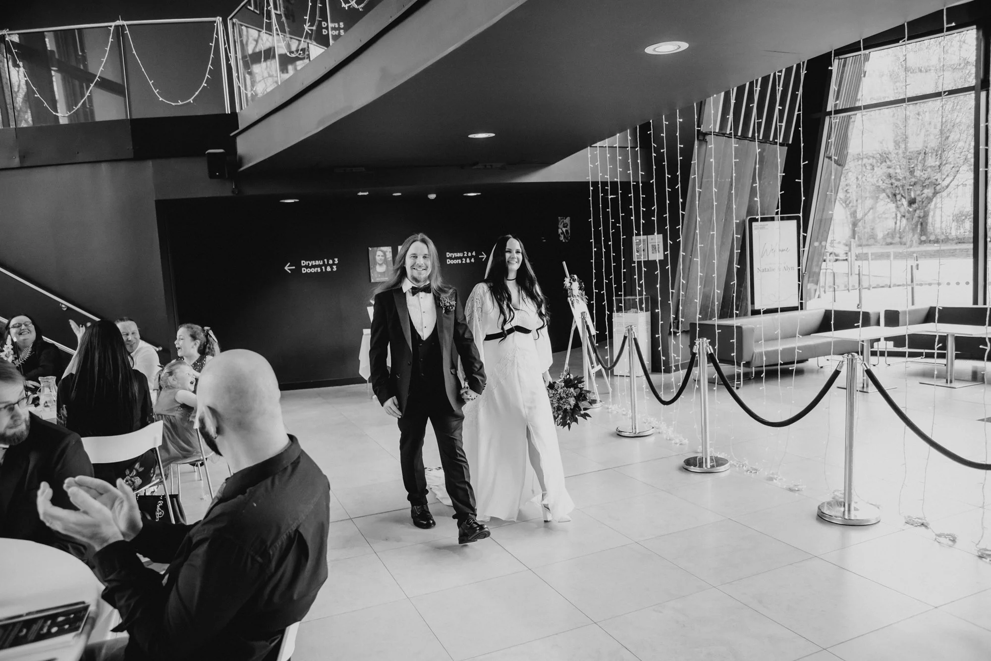 A wedding celebration with a couple walking hand in hand. The groom is dressed in a tuxedo, and the bride is in a white wedding gown holding a bouquet. Guests are seated at tables, clapping and smiling. The decor includes string lights and a sign in 