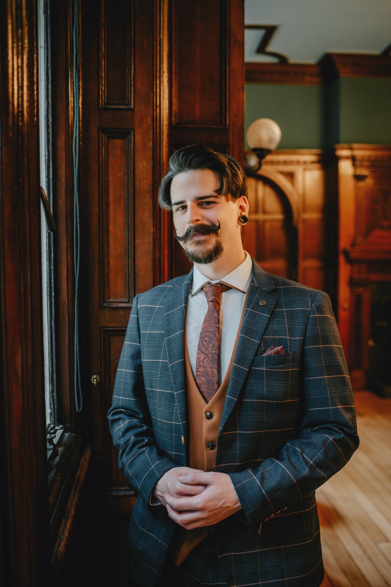 A man in a plaid suit and patterned tie stands indoors in front of wooden paneling, smiling at the camera.