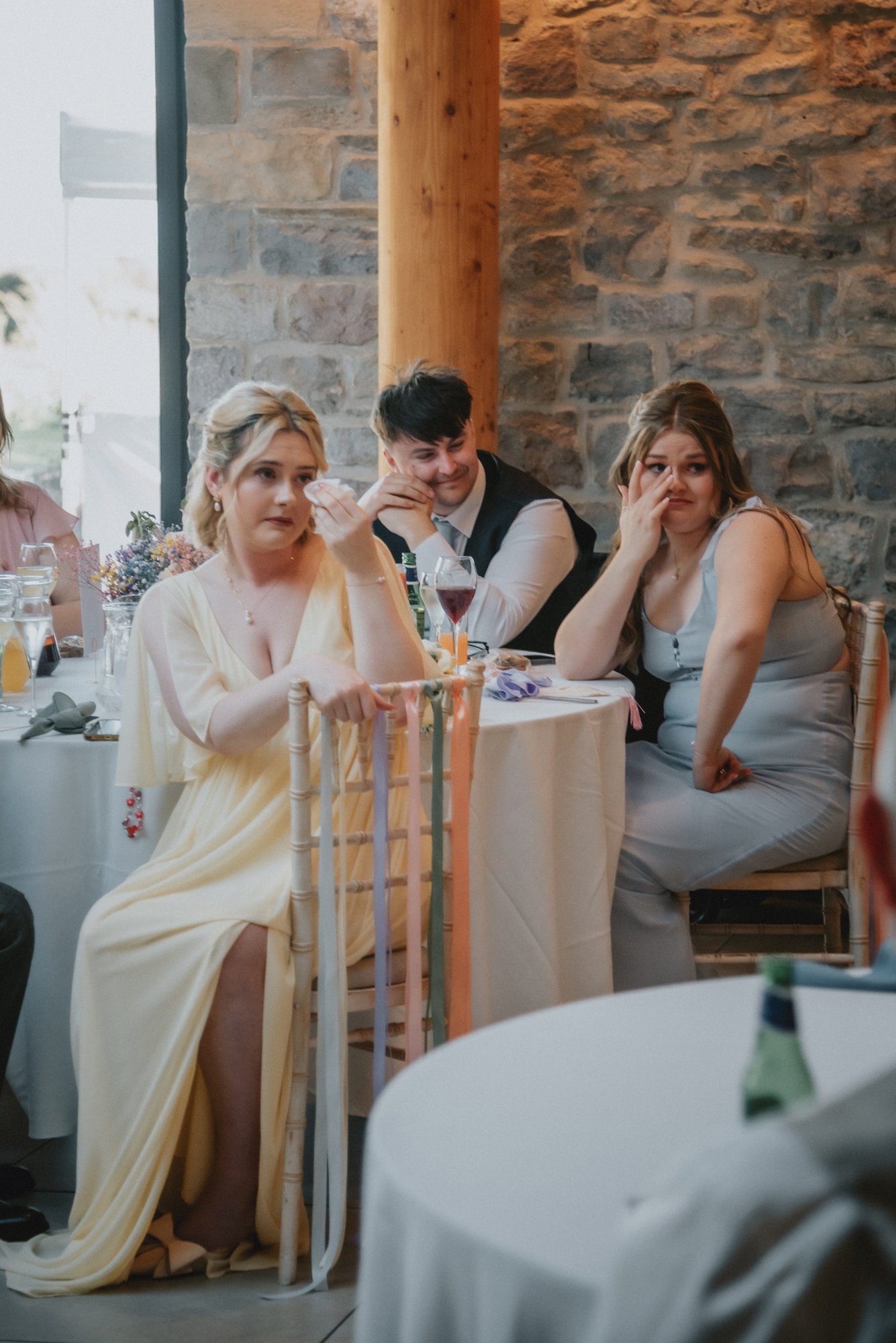 People sitting at a decorated table during a celebration, with a woman in a yellow dress, a man in a vest, and a woman in a gray dress, showing emotions of sadness or concern.