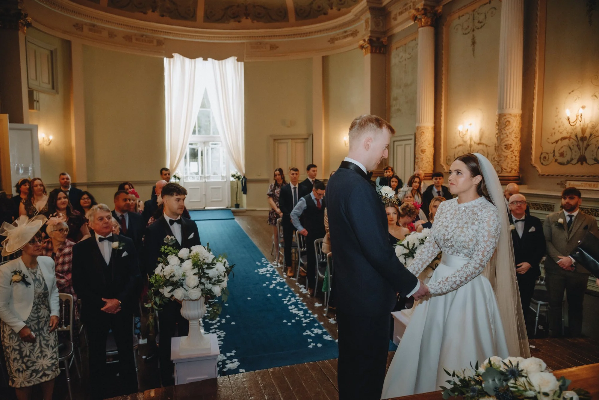 A wedding ceremony inside a grand, ornate hall with the bride and groom holding hands facing each other. Guests seated on either side of the aisle, paying attention to the couple. The hall has tall windows, white curtains, and decorative walls with g
