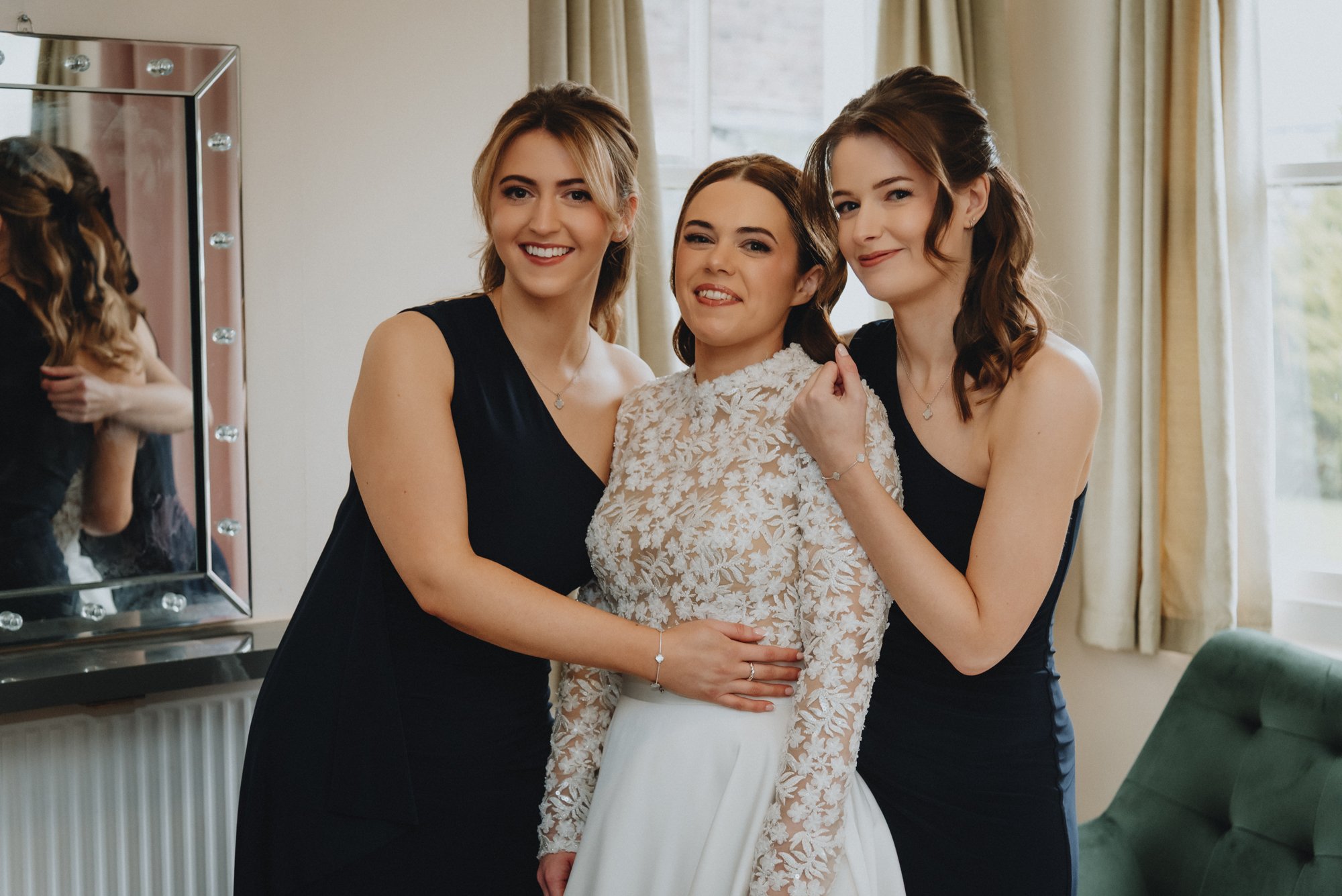 Three women smiling and posing together indoors, with one in a white lace dress, possibly a wedding dress, and two in black dresses, on a bright day with a window and green armchair in the background.