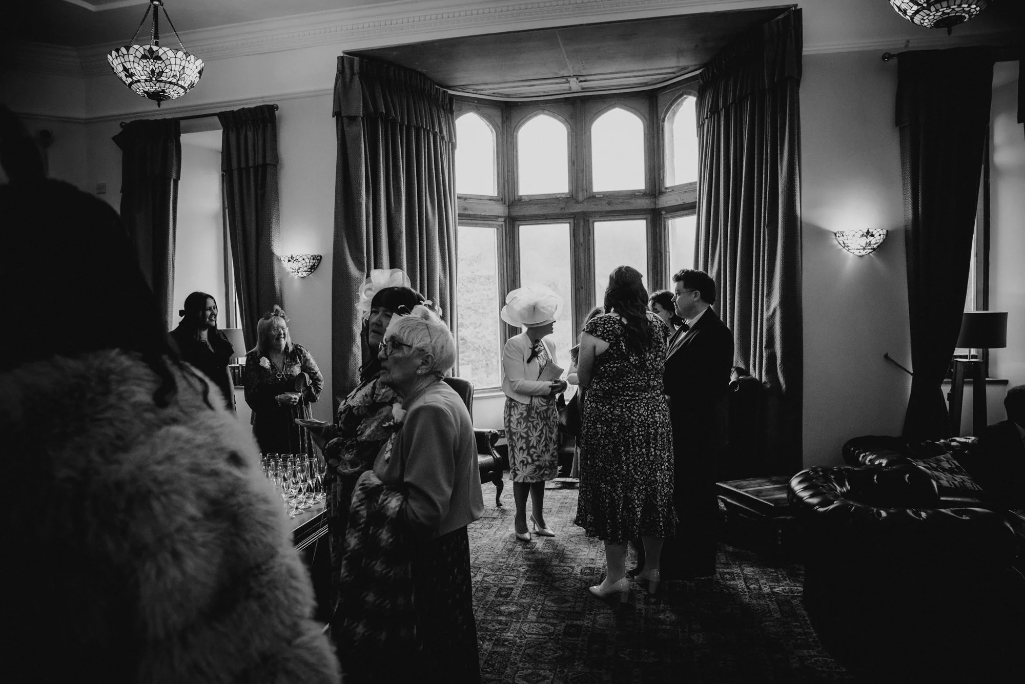 Black and white photo of a social gathering in a room with large window and heavy curtains. Several people are talking and holding drinks, some dressed in formal and vintage attire, including women in hats. The room has ornate light fixtures and a pa