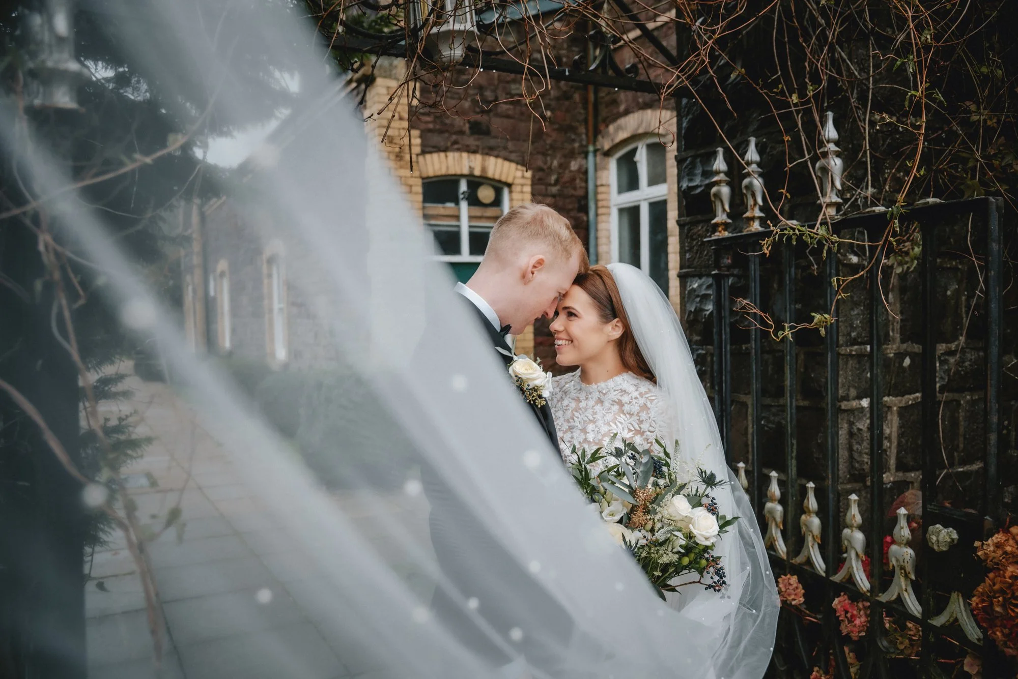 Bride and groom standing close together, smiling and touching foreheads outside a brick building with iron fence and leafless vines, bride holding bouquet of flowers, woman in wedding dress and veil, man in tuxedo.