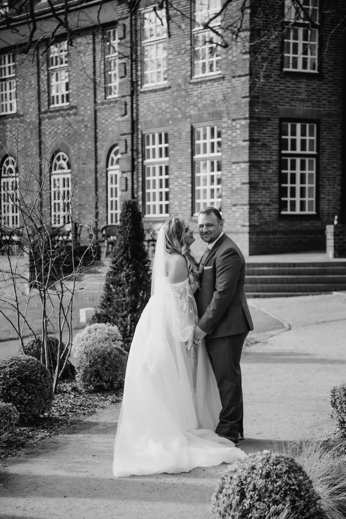A bride and groom in wedding attire holding hands and smiling outside in front of a brick building with large windows, surrounded by bushes.
