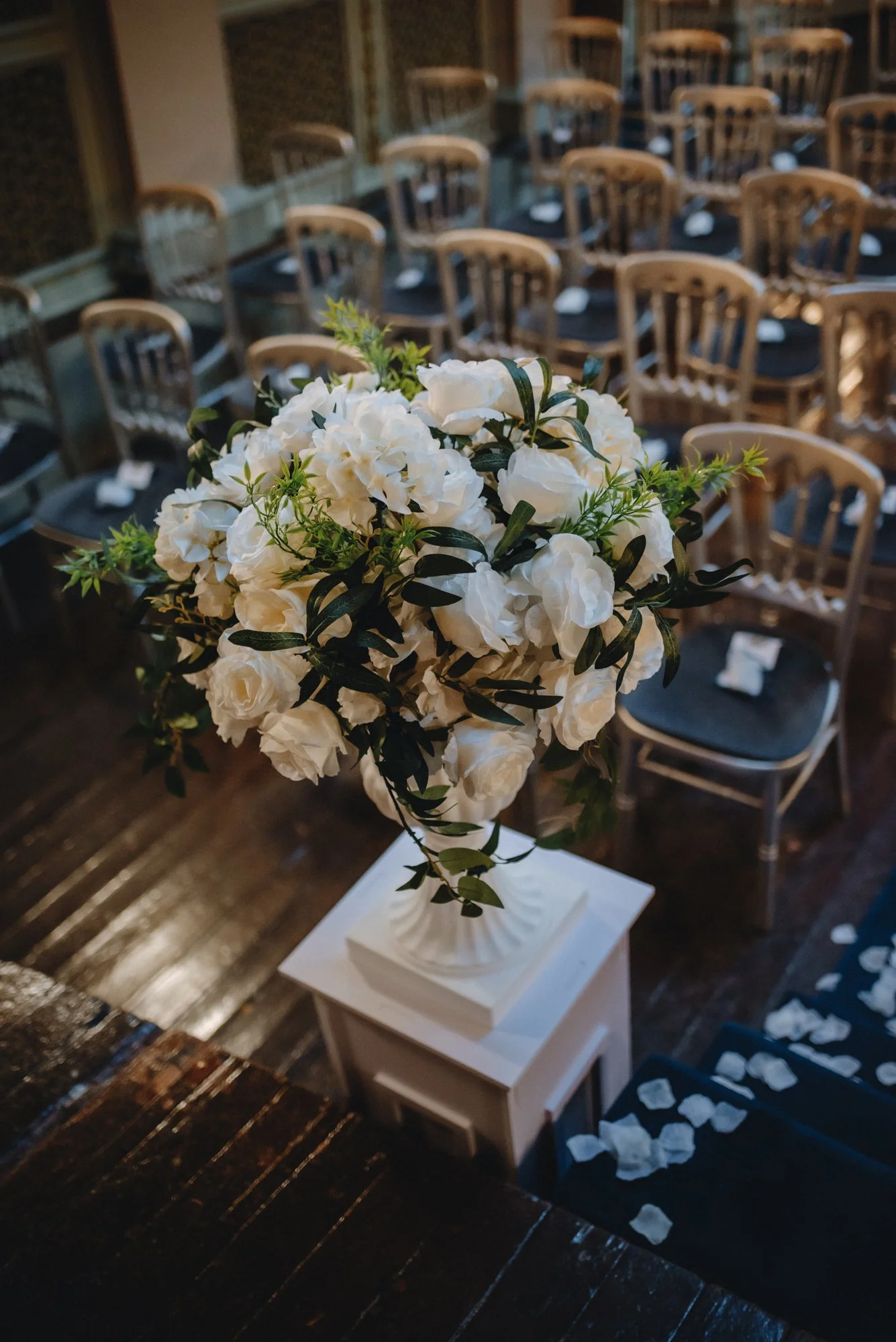 A large bouquet of white flowers and greenery in a white vase on a small white pedestal, in front of rows of wooden chairs with black seats, inside a decorated venue.