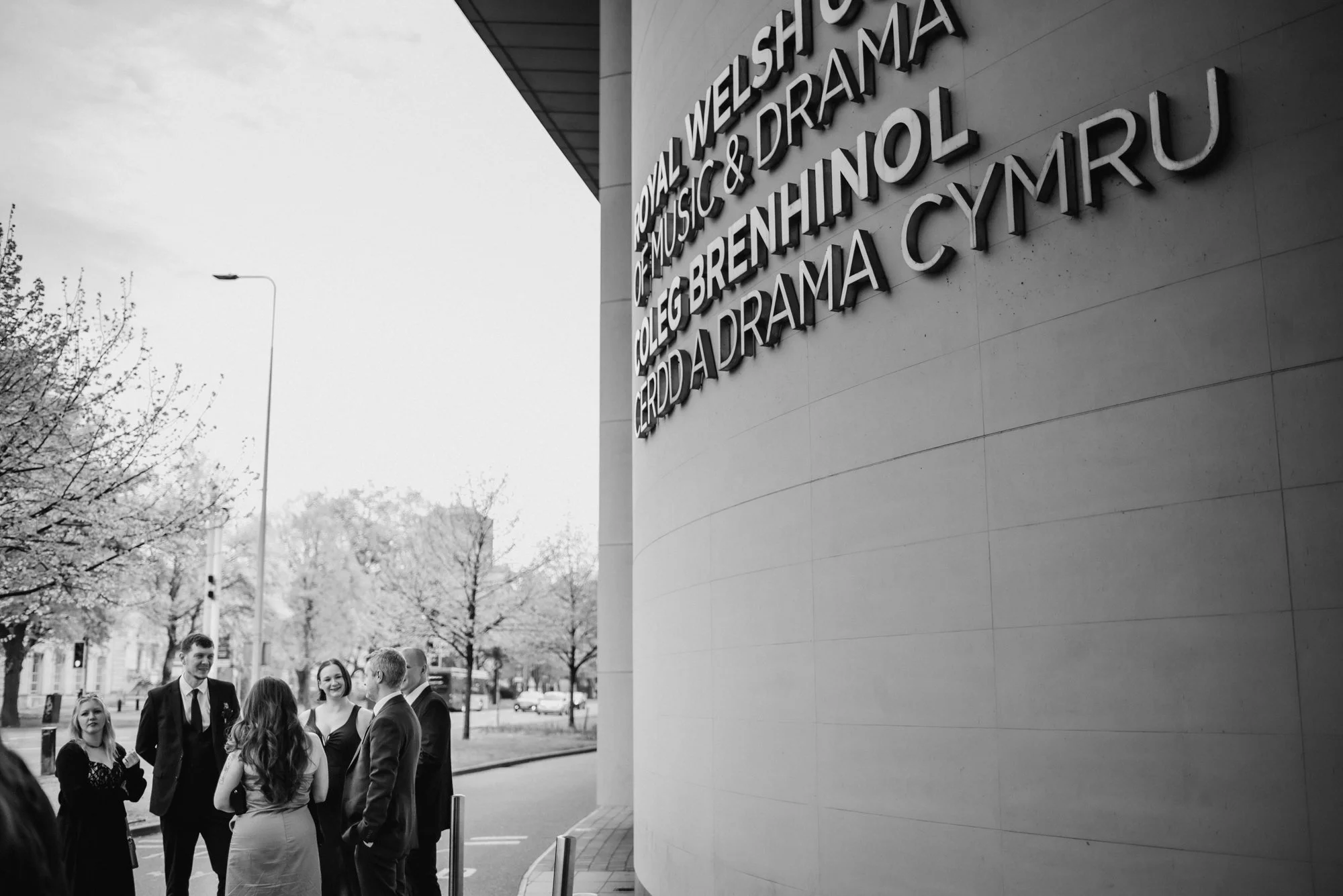 A group of people in formal attire standing outside the Royal Welsh College of Music & Drama in Wales, with building signage on a curved wall and trees in the background.