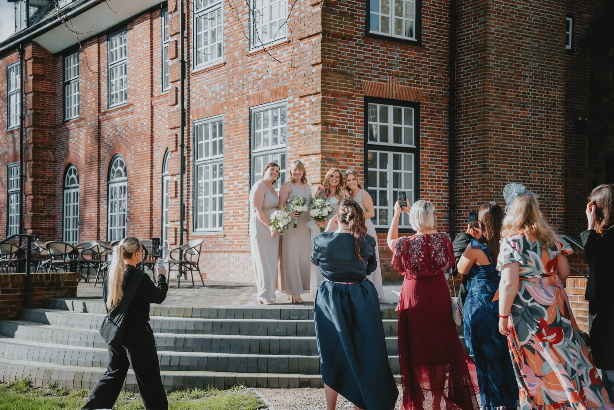 Women in beige dresses holding bouquets standing on a brick building's outdoor steps, surrounded by people taking pictures at a wedding or celebration.