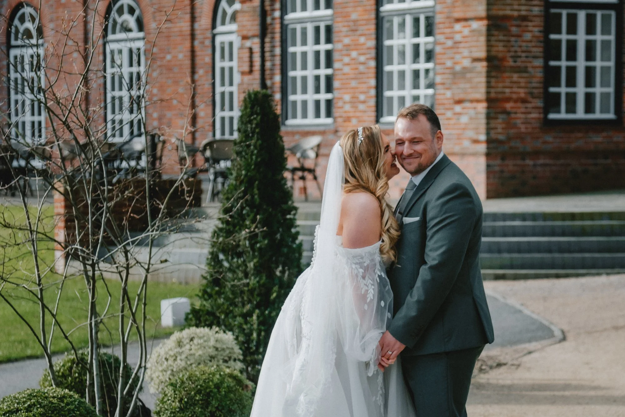 A bride and groom standing outside a brick building, smiling and holding hands, during their wedding celebration.