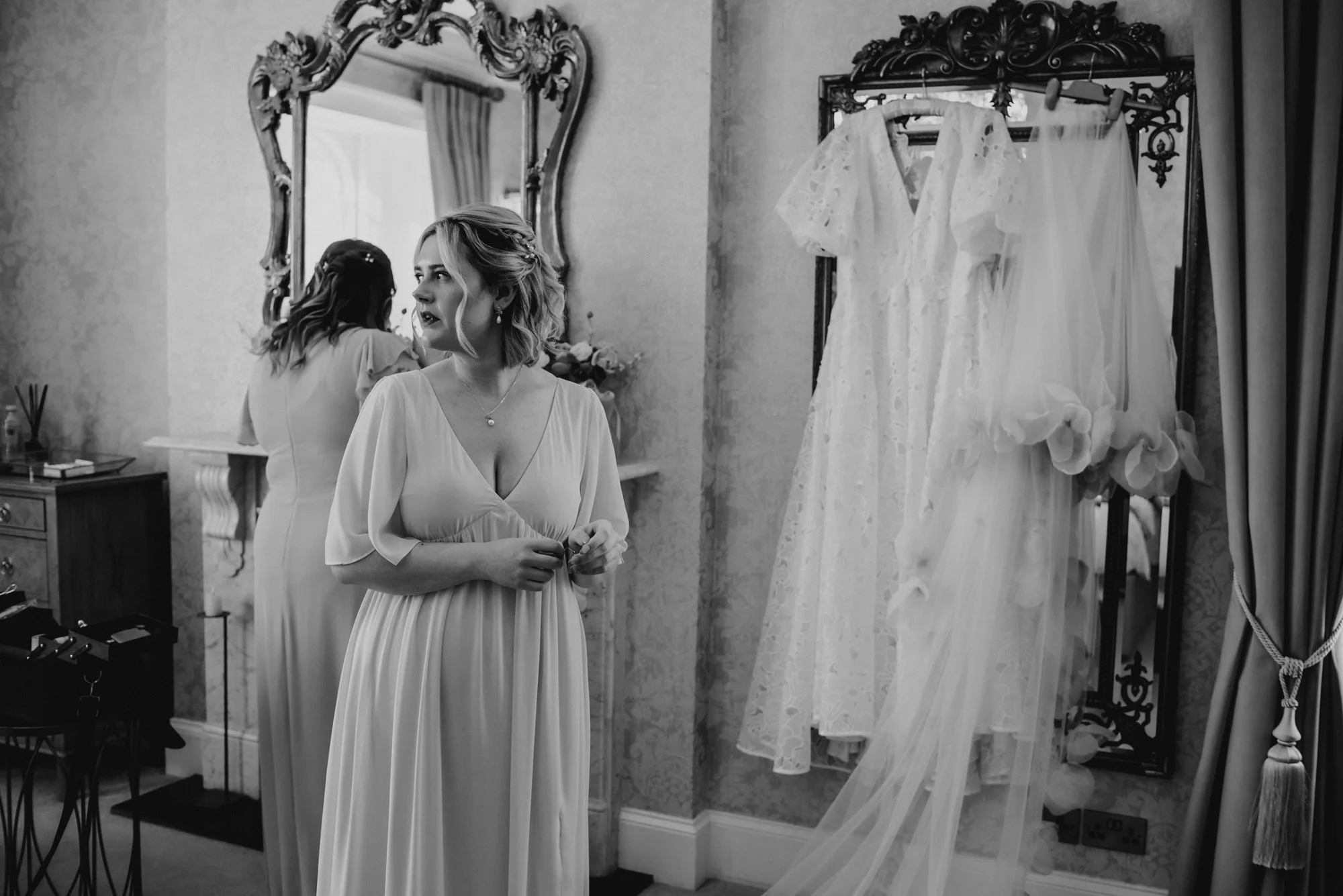 A woman in a light-colored dress standing in a room with ornate mirrors, looking to her left. Behind her, another woman wears a similar dress, with her back to the camera, and a table with small items. On the right side, a stand holds three hanging w
