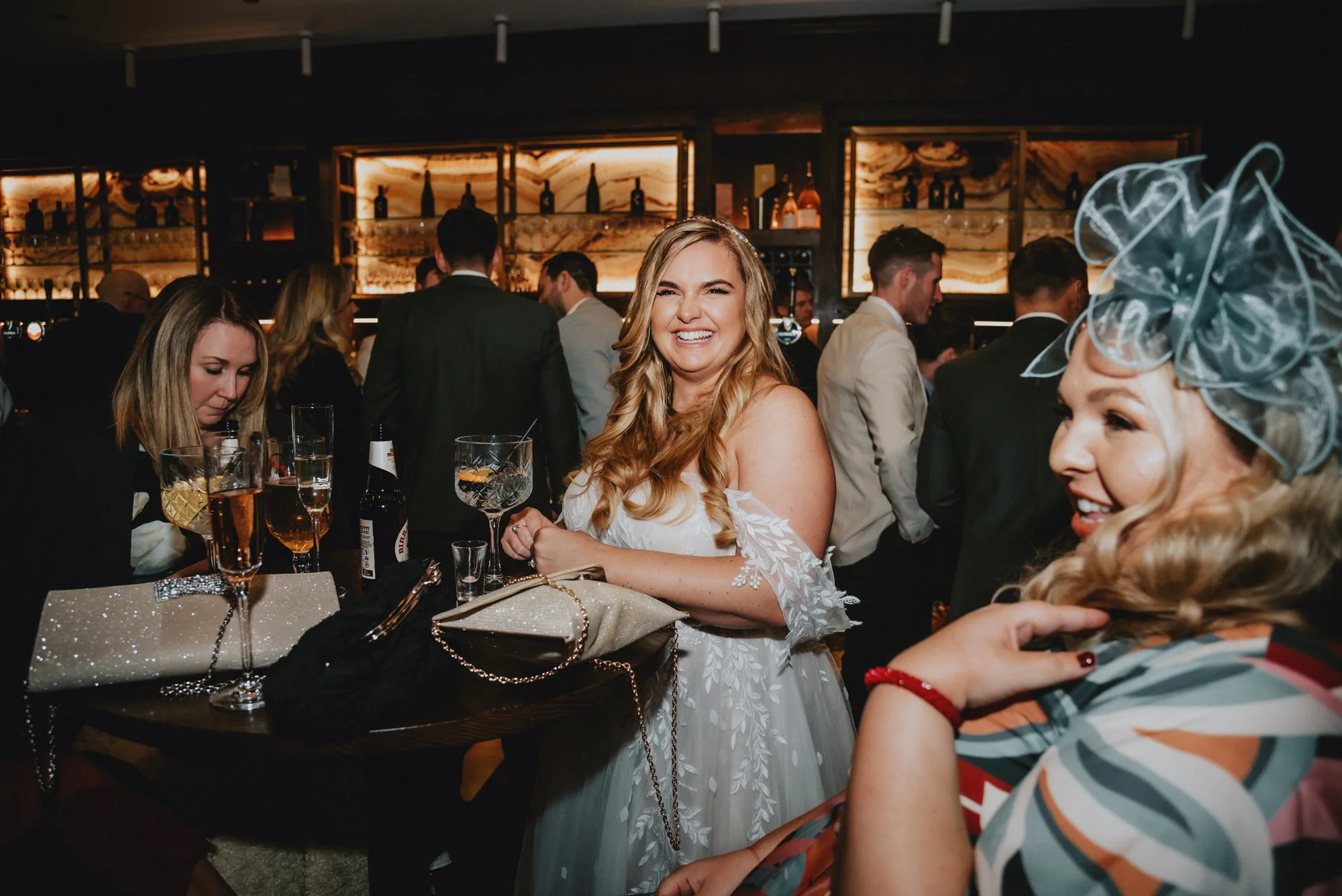 A woman in a white dress smiling and socializing at a crowded event in a dimly lit venue with a bar in the background.