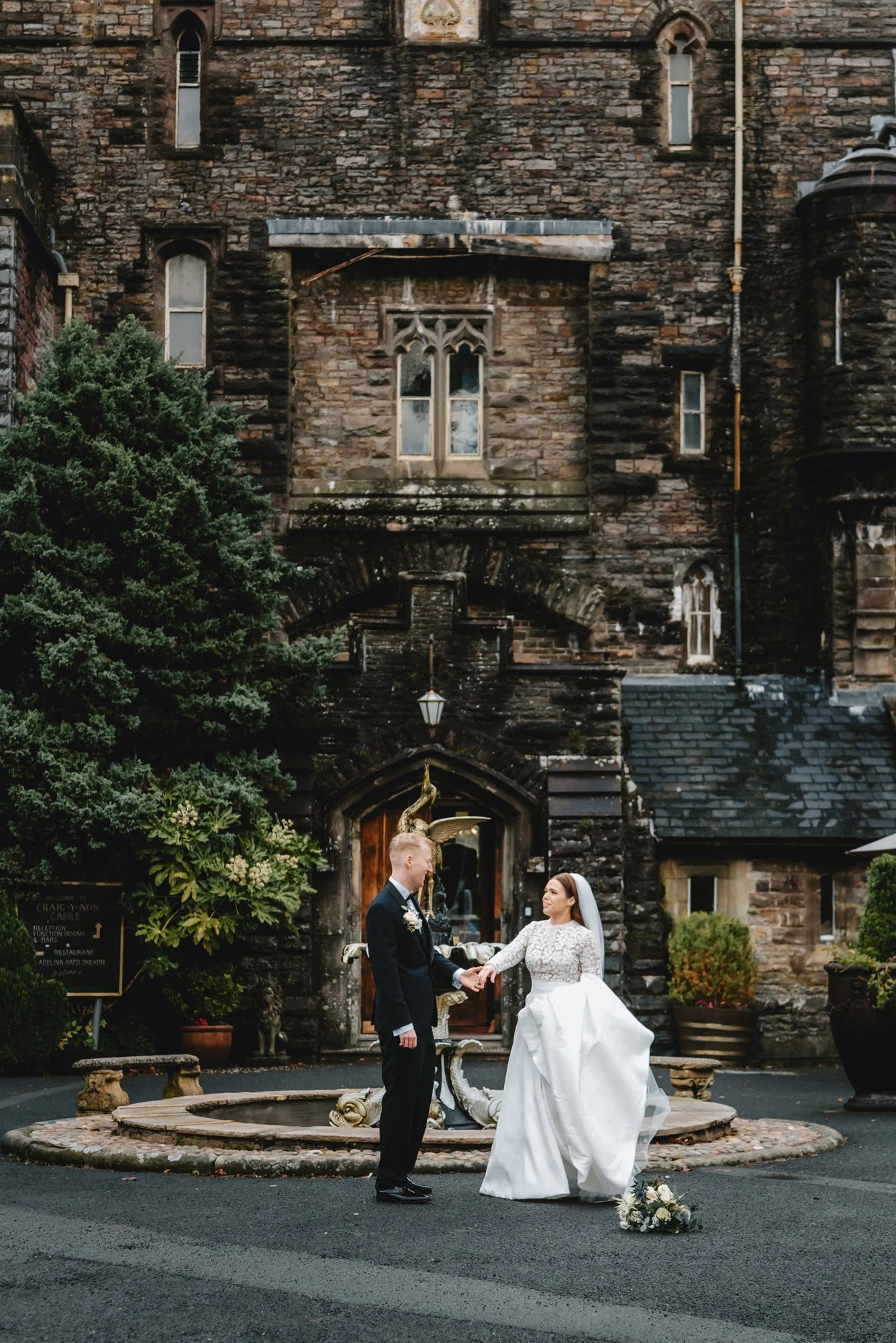 A bride and groom holding hands in front of a historic stone building with gothic windows, a fountain, and greenery outside during their wedding ceremony.