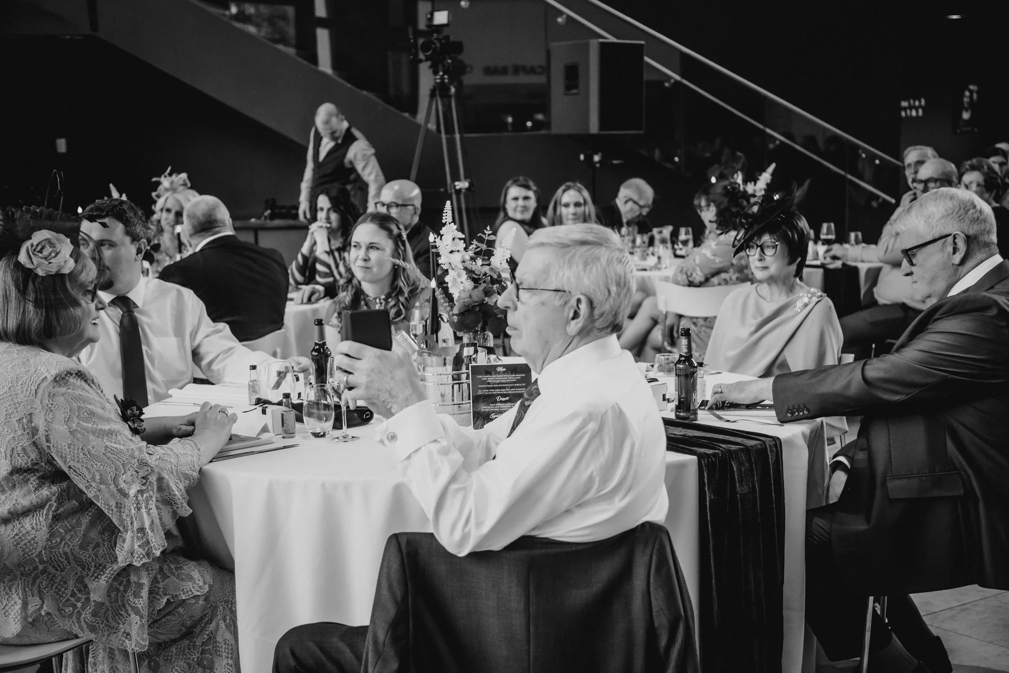 People seated at banquet tables during a formal event, with some taking photos, in an indoor venue with a staircase and cameras in the background.