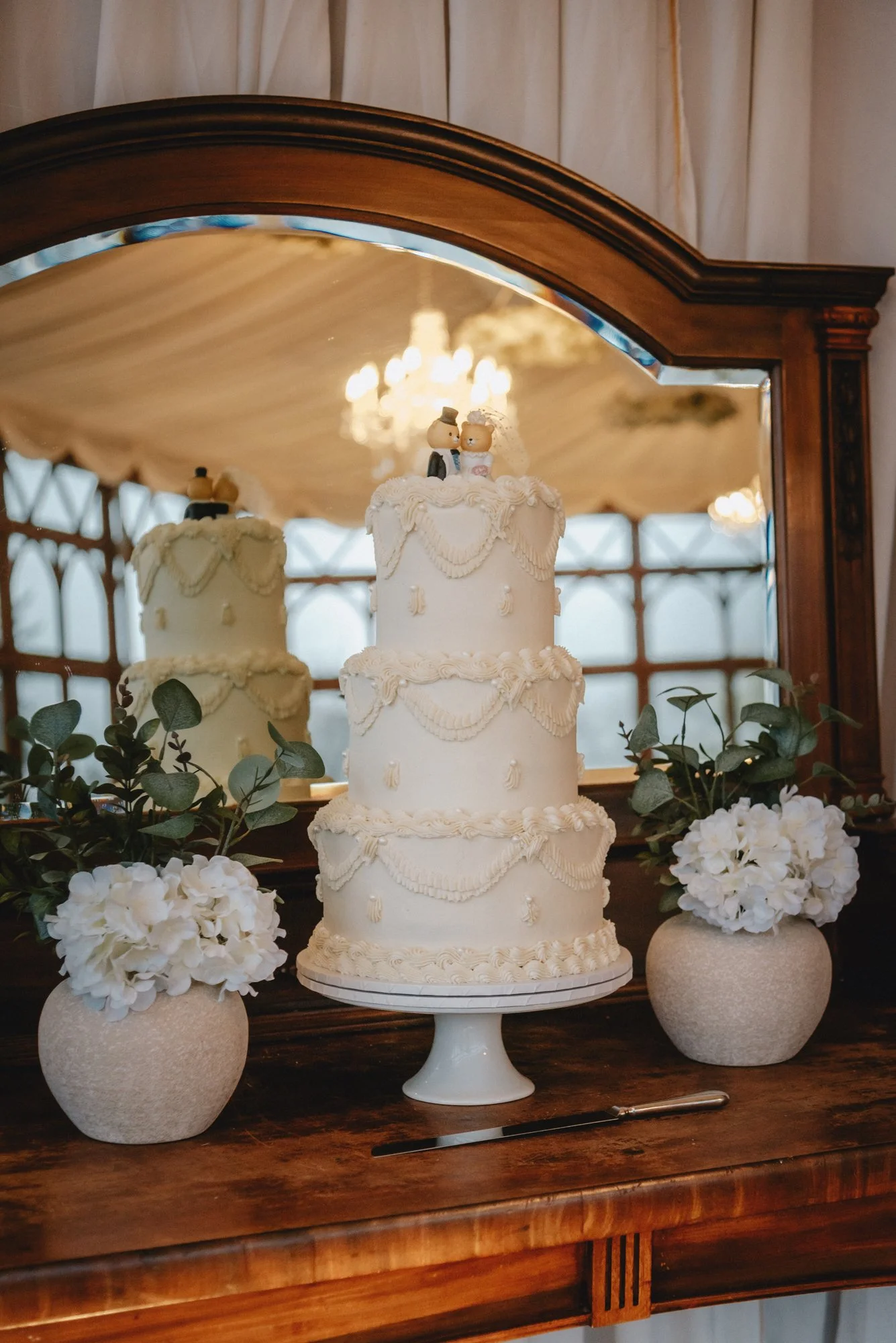 A tiered wedding cake decorated with white icing and ornate piping, topped with a bride and groom figurine, flanked by white hydrangea arrangements in textured pots, with a large mirror reflecting the cake and a chandelier overhead.