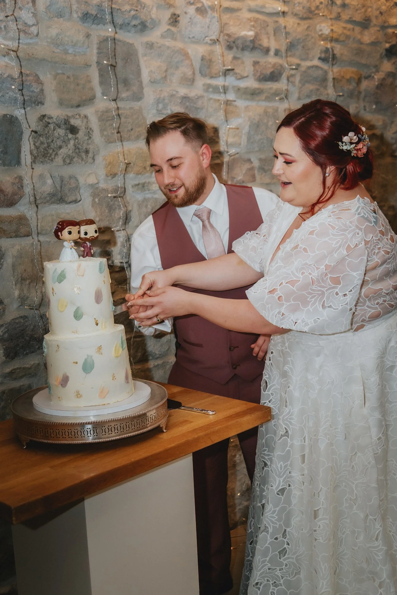 A bride and groom cutting a wedding cake together, adorned with whimsical figurines on top, against a stone wall background.