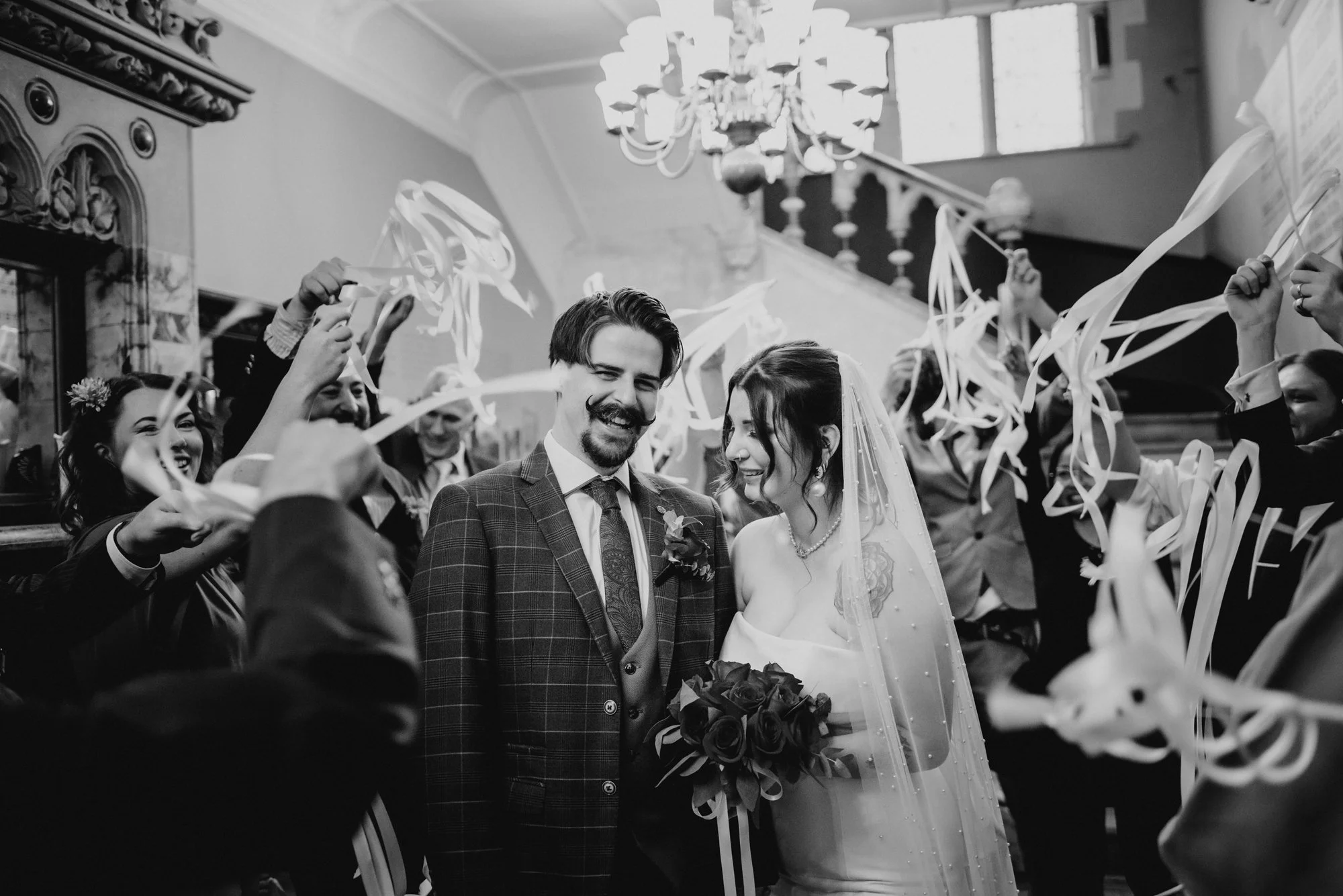 A bride and groom smiling at each other while standing in the center amid friends and family at a wedding reception, with guests waving streamers around them in a decorated hall with a chandelier.