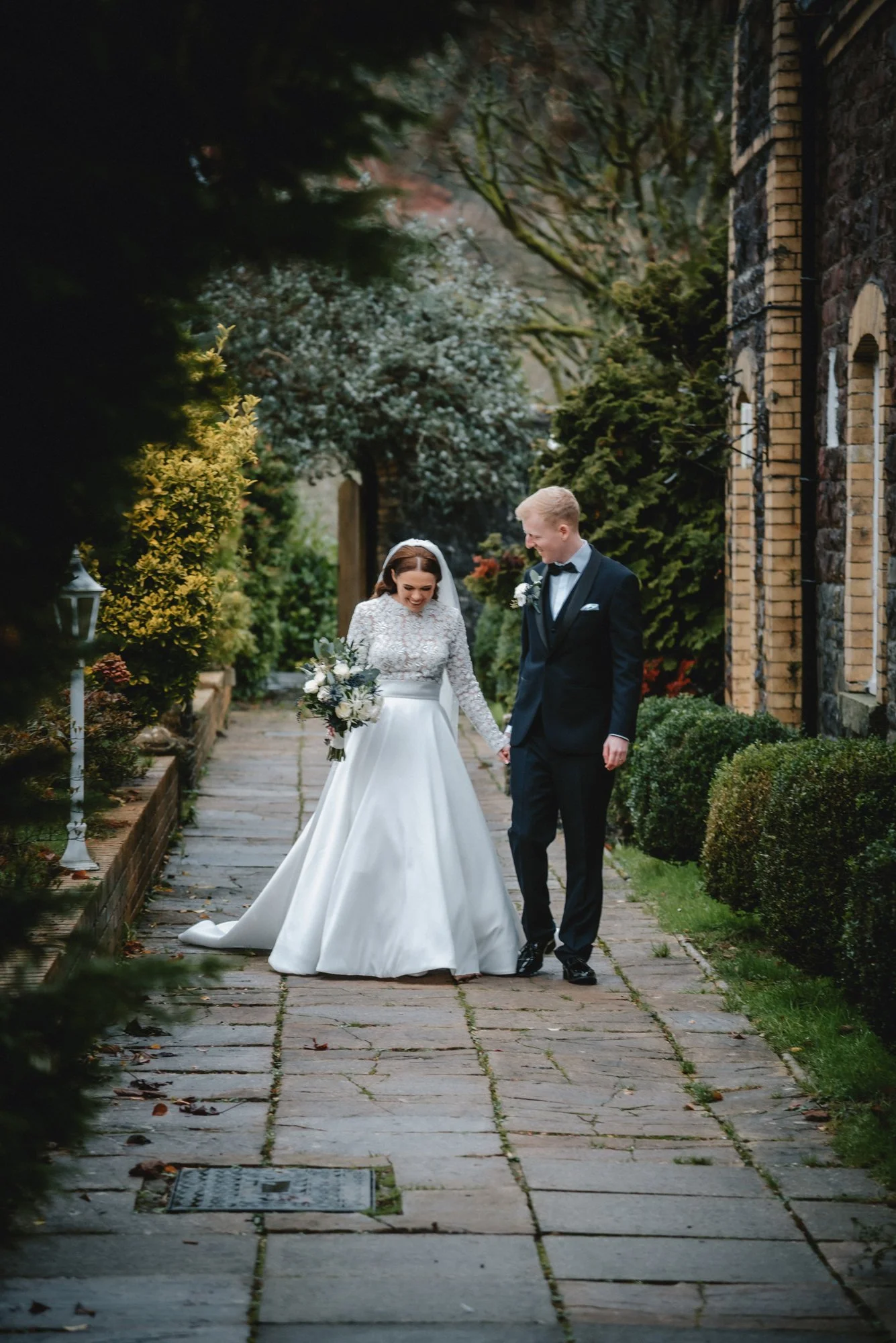 Bride and groom walking hand in hand on a stone pathway outside, surrounded by greenery and a stone building in the background, during their wedding.