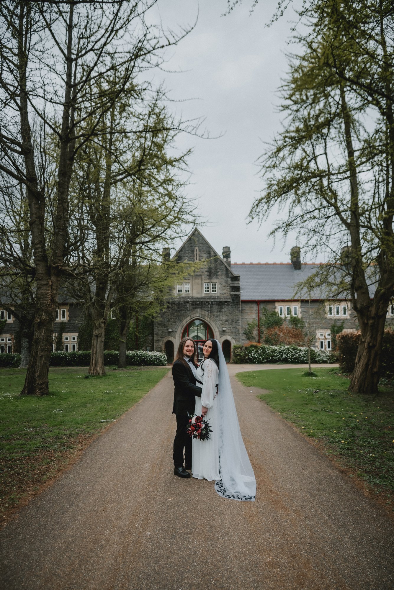 A bride and groom standing on a pathway in front of a large stone building with Gothic architecture, surrounded by trees and greenery.