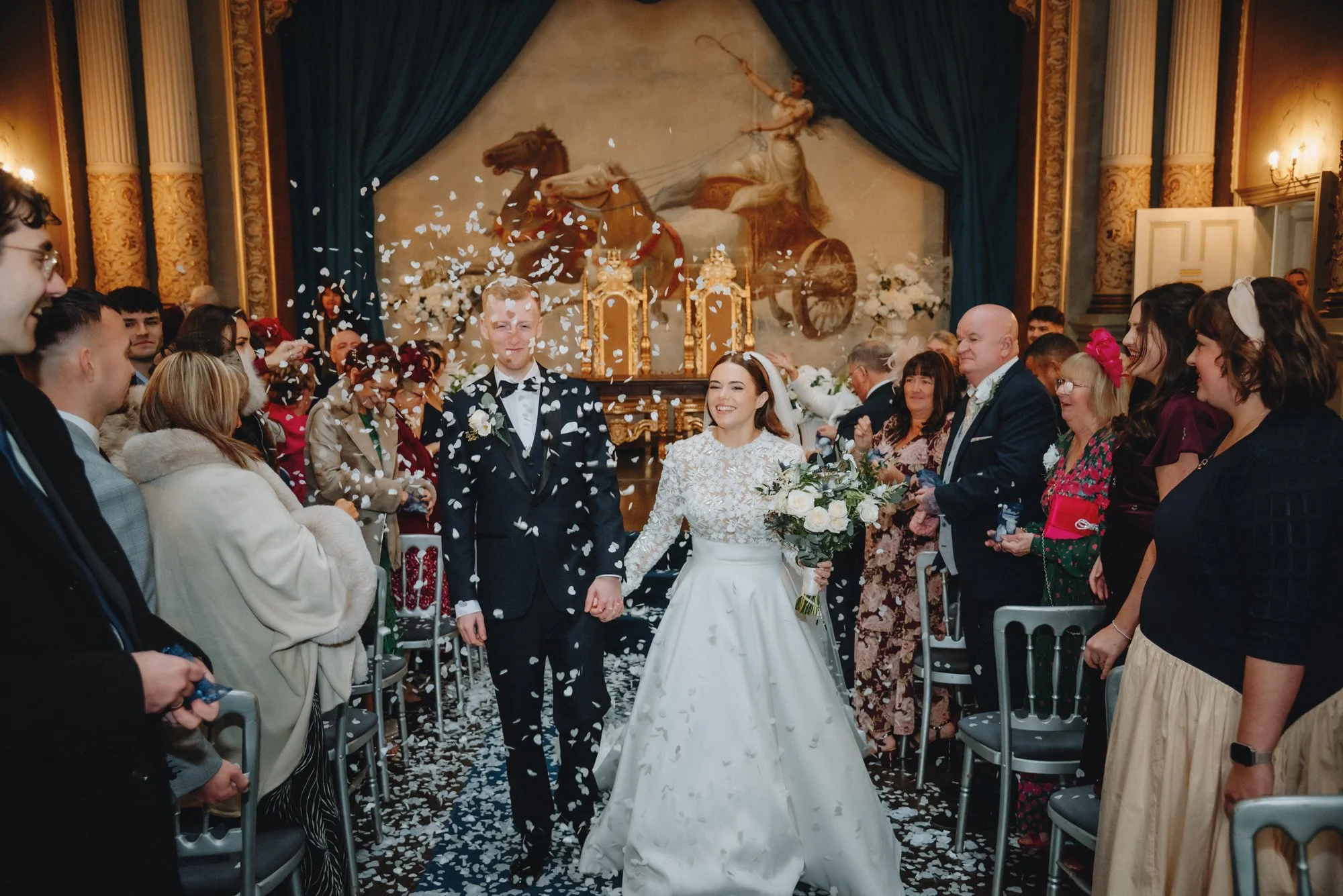 A bride and groom walk down the aisle holding hands at their wedding, surrounded by guests throwing flower petals and smiling.