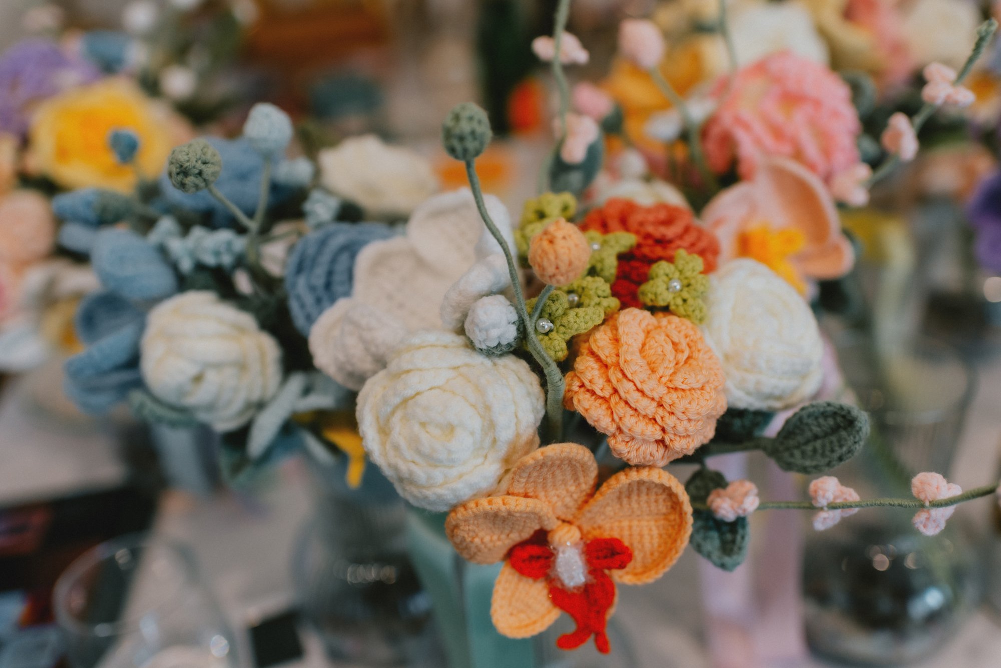 Close-up of a bouquet of crocheted flowers in various colors including white, orange, peach, green, and purple, arranged in a vase.