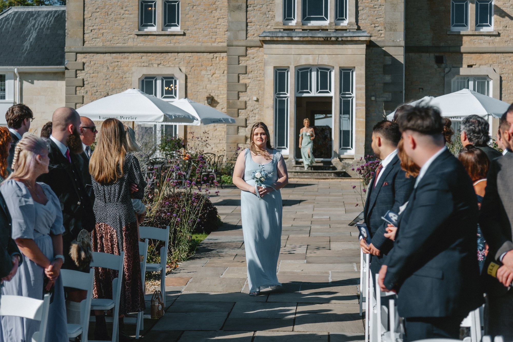 A woman in a light blue dress holding a bouquet walking down an aisle outdoors at a wedding ceremony, with guests seated on either side and a large stone building in the background.