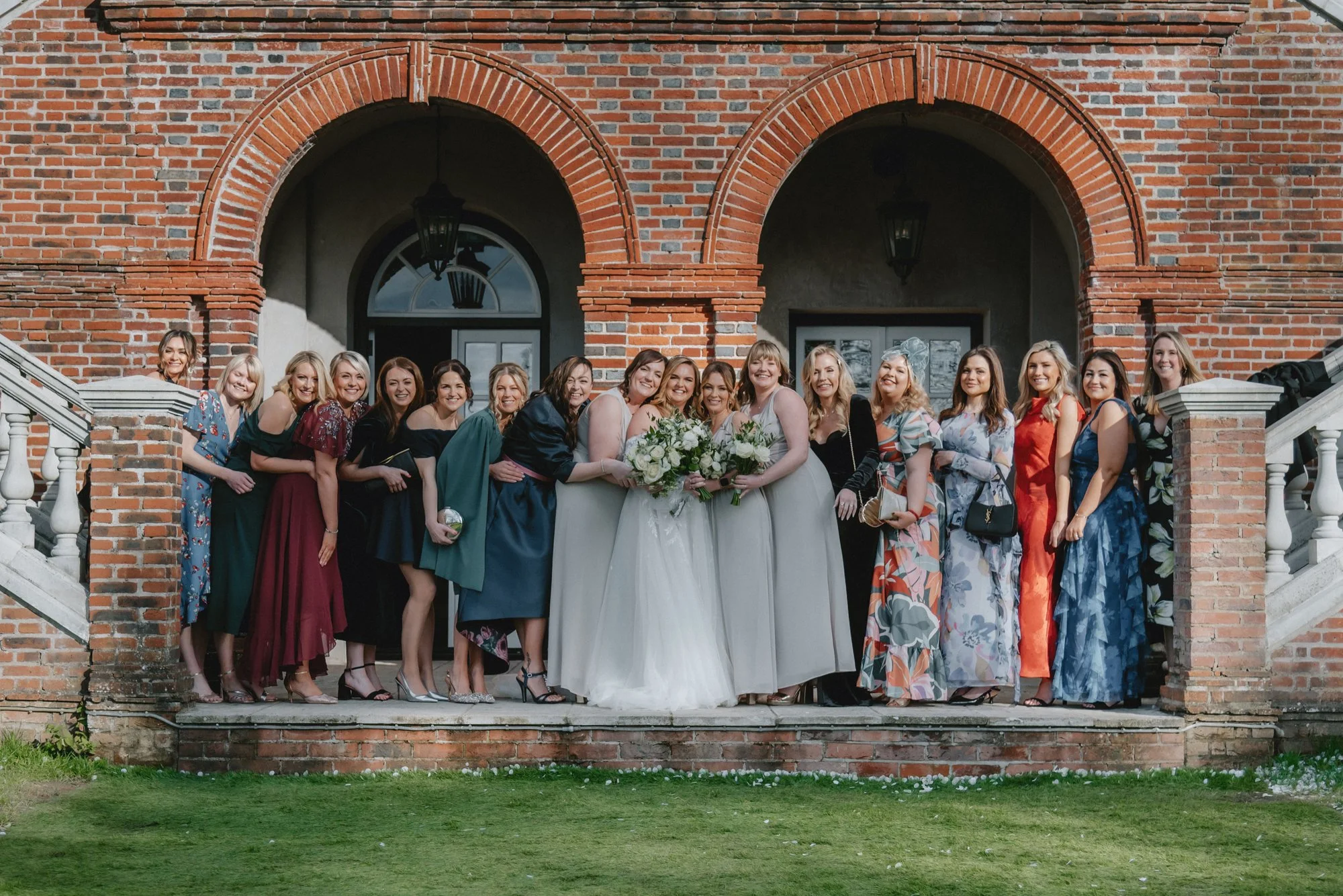 A group of women, including two brides in wedding dresses holding bouquets, standing on steps in front of a brick building with arches and windows, celebrating a wedding.