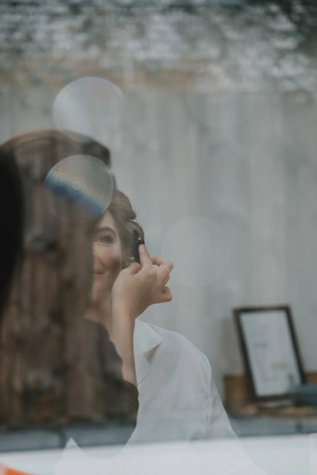 A woman is getting her makeup done, seen through a reflective surface with bokeh lights in the background.