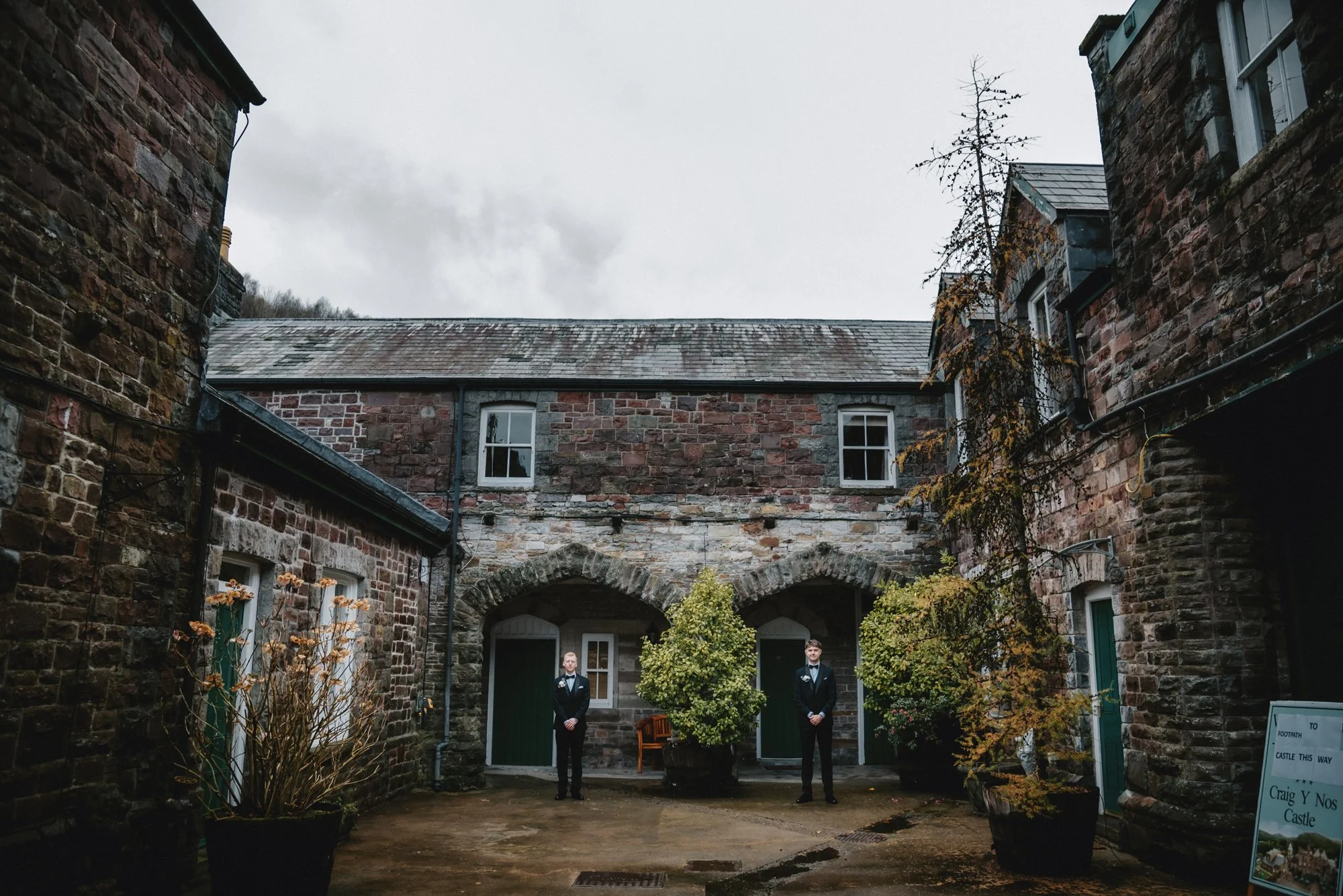 Two men in black suits standing in a stone courtyard with large potted plants and an old brick building in the background.