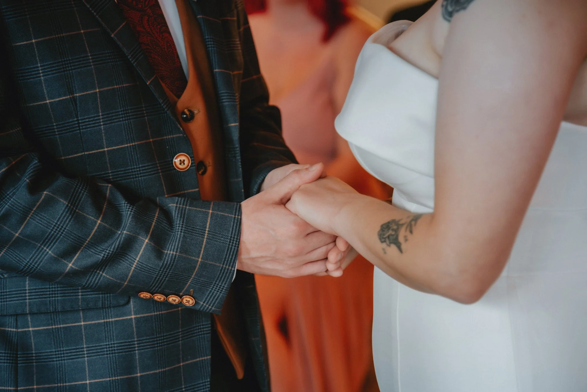 A couple holding hands, with one wearing a dark plaid suit and the other wearing a white dress, possibly at a wedding.