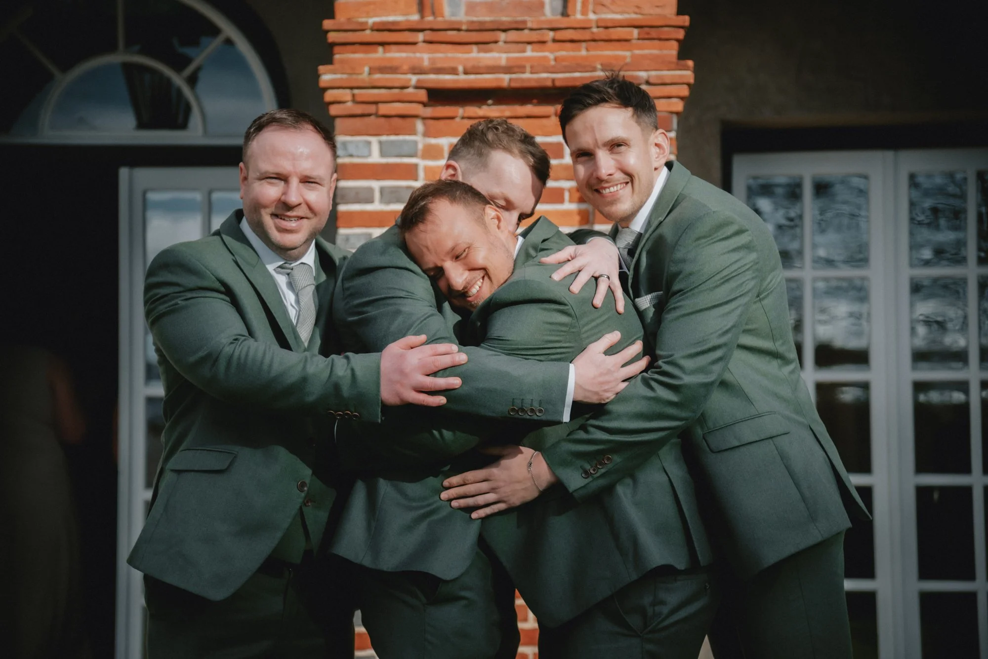 Five men in suits hugging and smiling together outside a building with brick and glass windows.