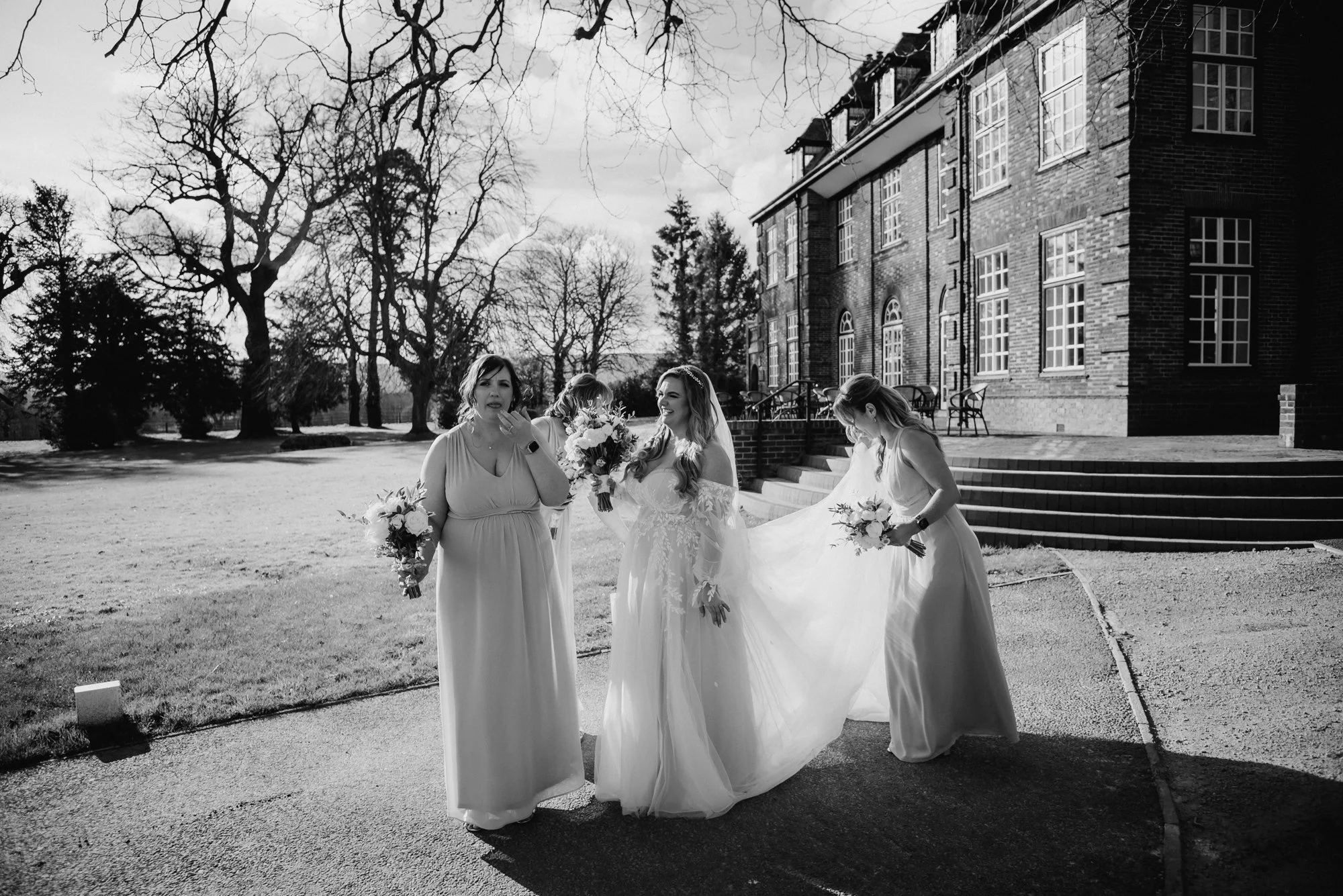 A group of women, one in a wedding dress, standing outdoors near a brick building with steps, holding bouquets, with leafless trees and a clear sky in the background.