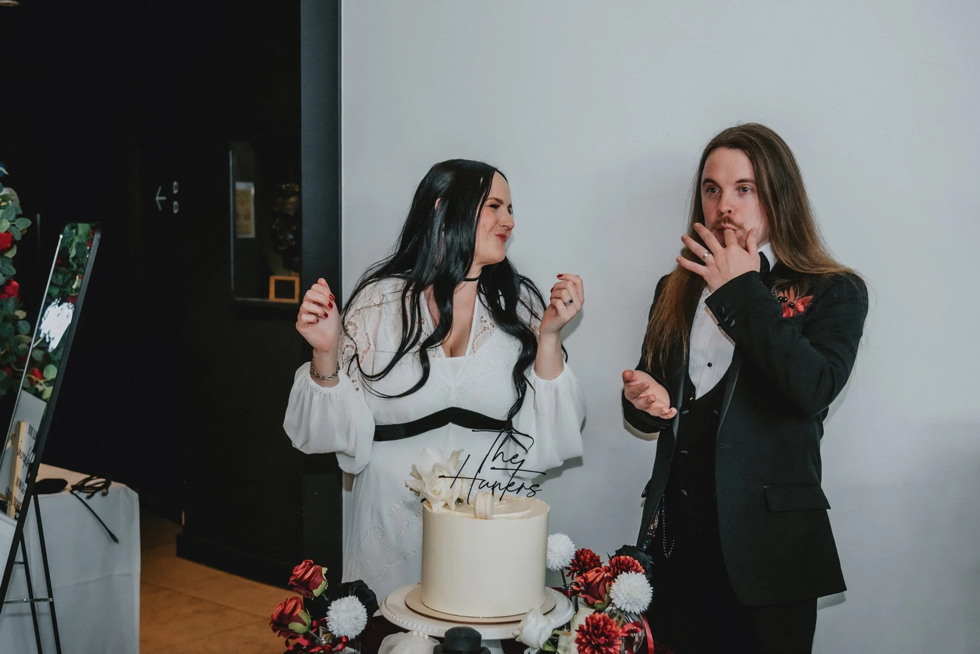 A woman and a man standing behind a wedding cake at a celebration. The woman is wearing a white dress, and the man is in a black tuxedo with a floral embroidery. The cake has a topper that says "The Harrpers" and is decorated with red, white, and bla