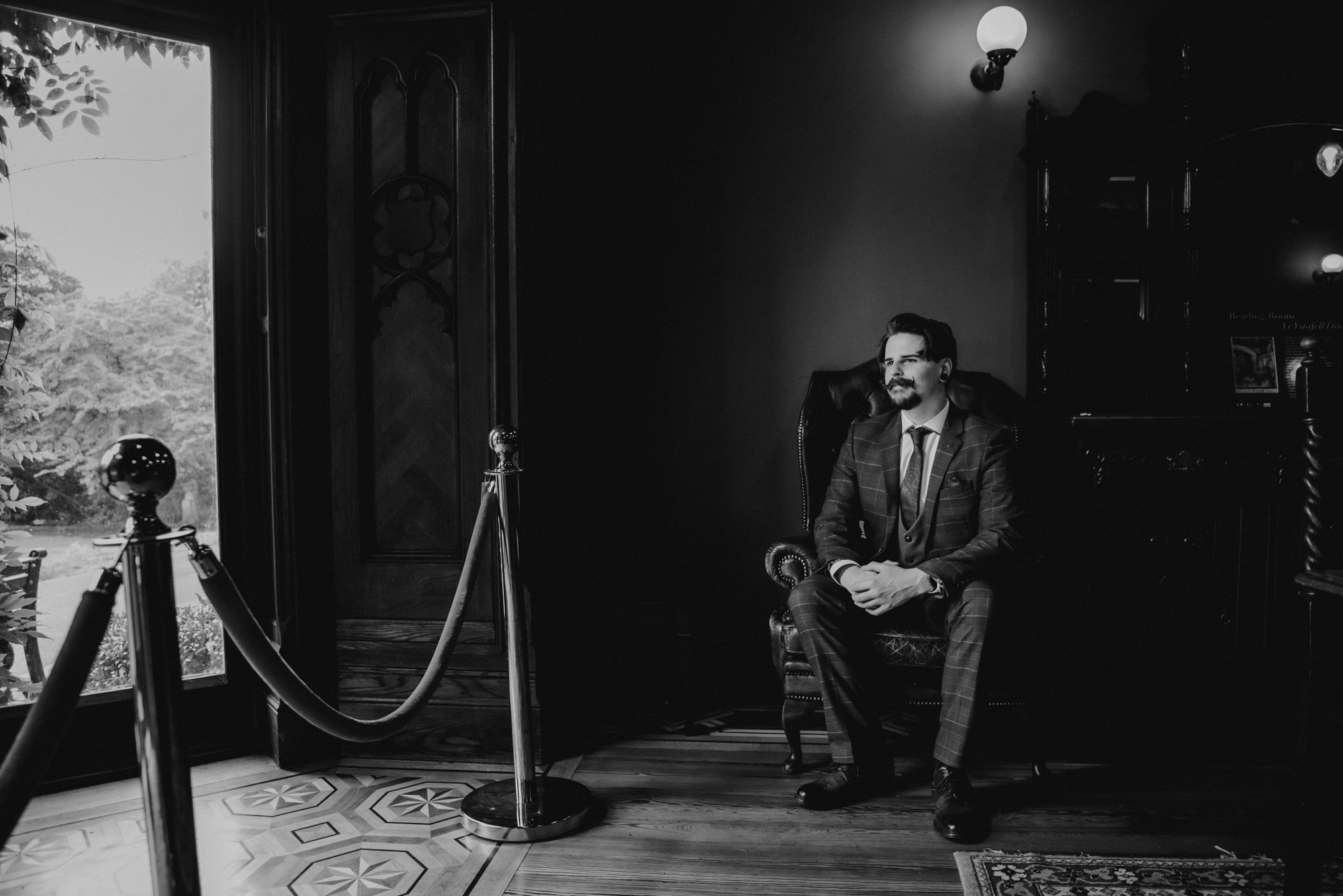 A man in a suit sitting on a vintage armchair inside a room with wooden floors and dark-paneled walls, illuminated by a wall-mounted light fixture. A large window with a velvet rope barrier is visible to the left, showing trees outside.