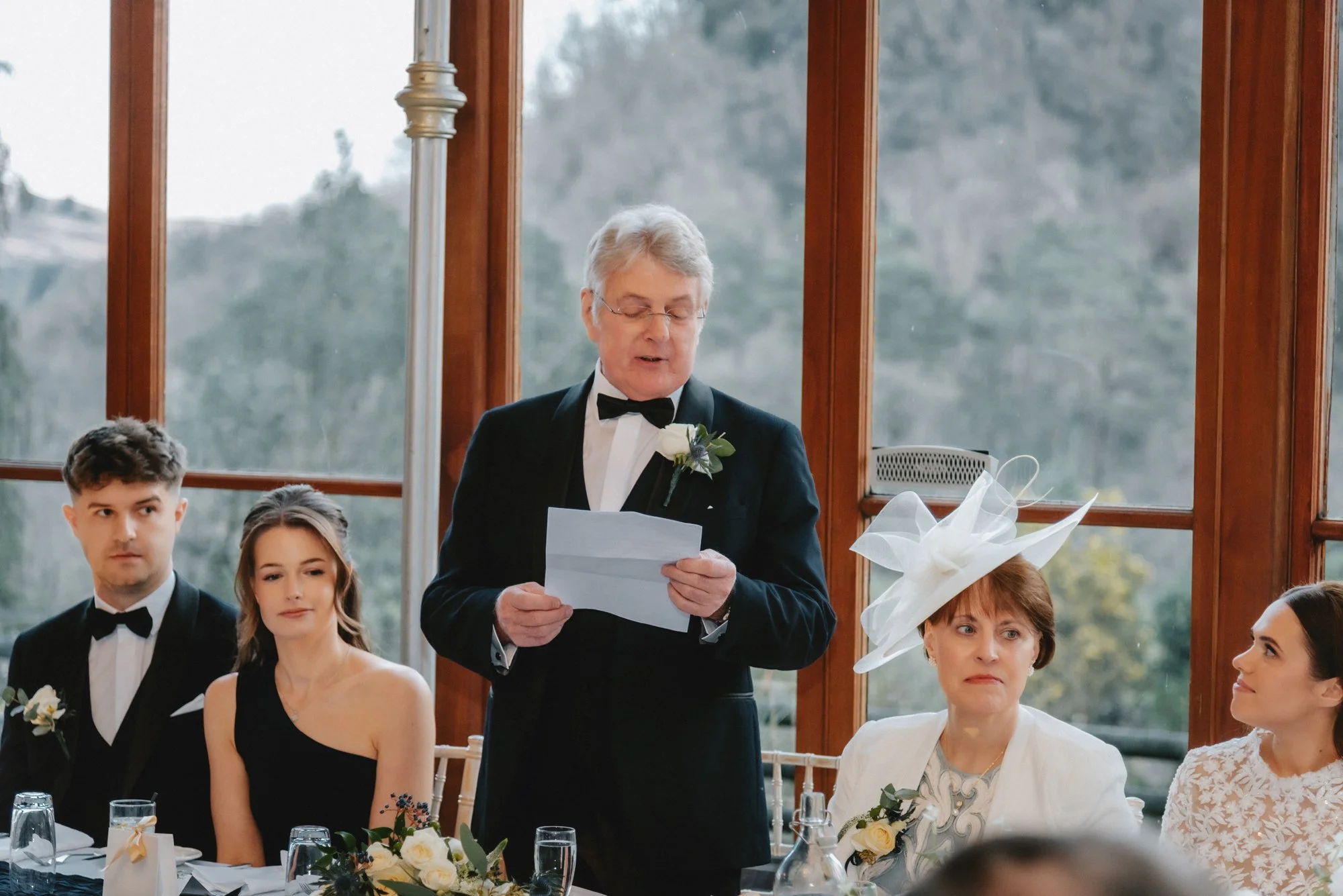 A man in a tuxedo giving a speech at a wedding reception, with women and men seated at a table decorated with flowers and glasses.