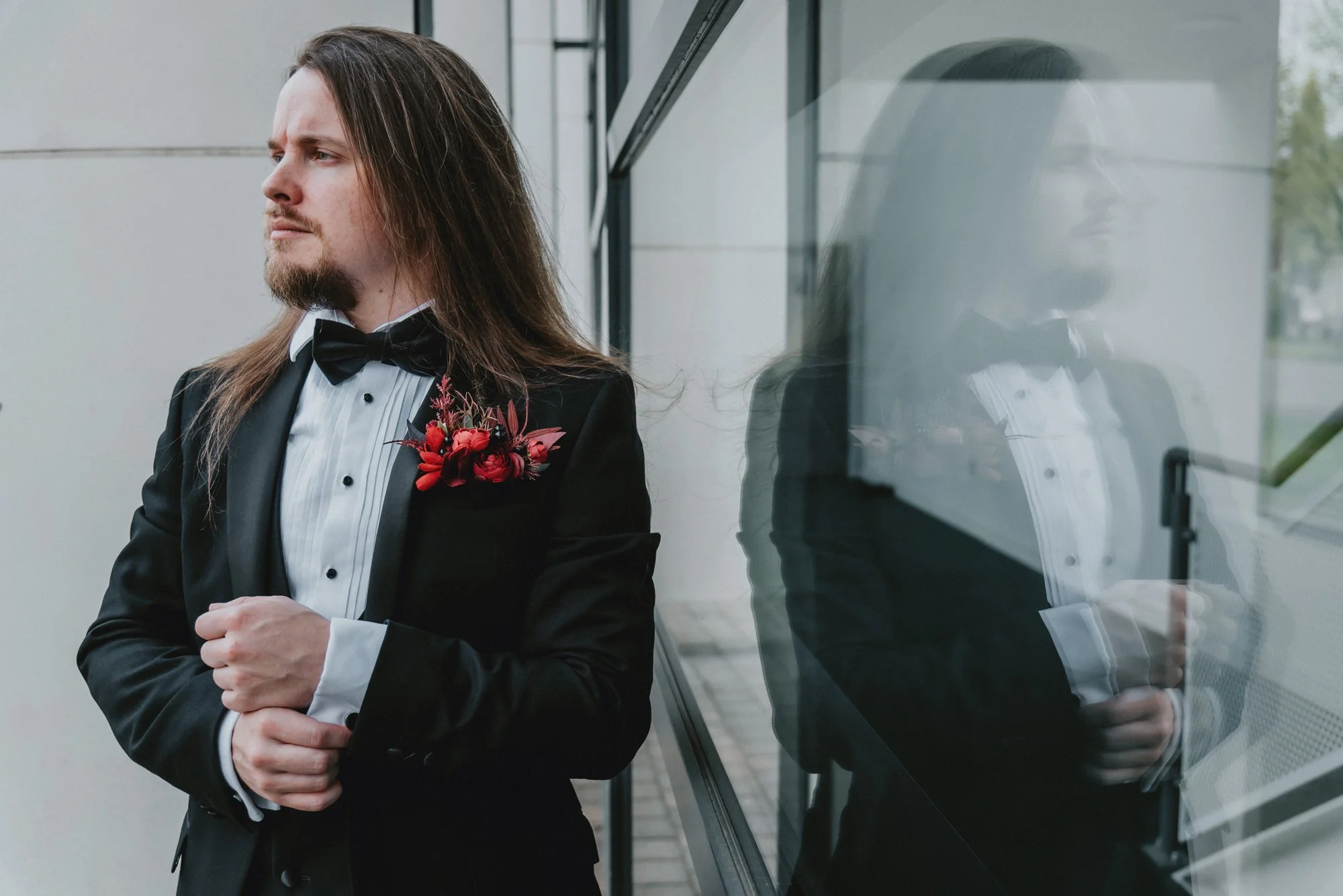 A man in a tuxedo with a flower boutonniere, standing outside near a glass wall, with his reflection visible on the glass.