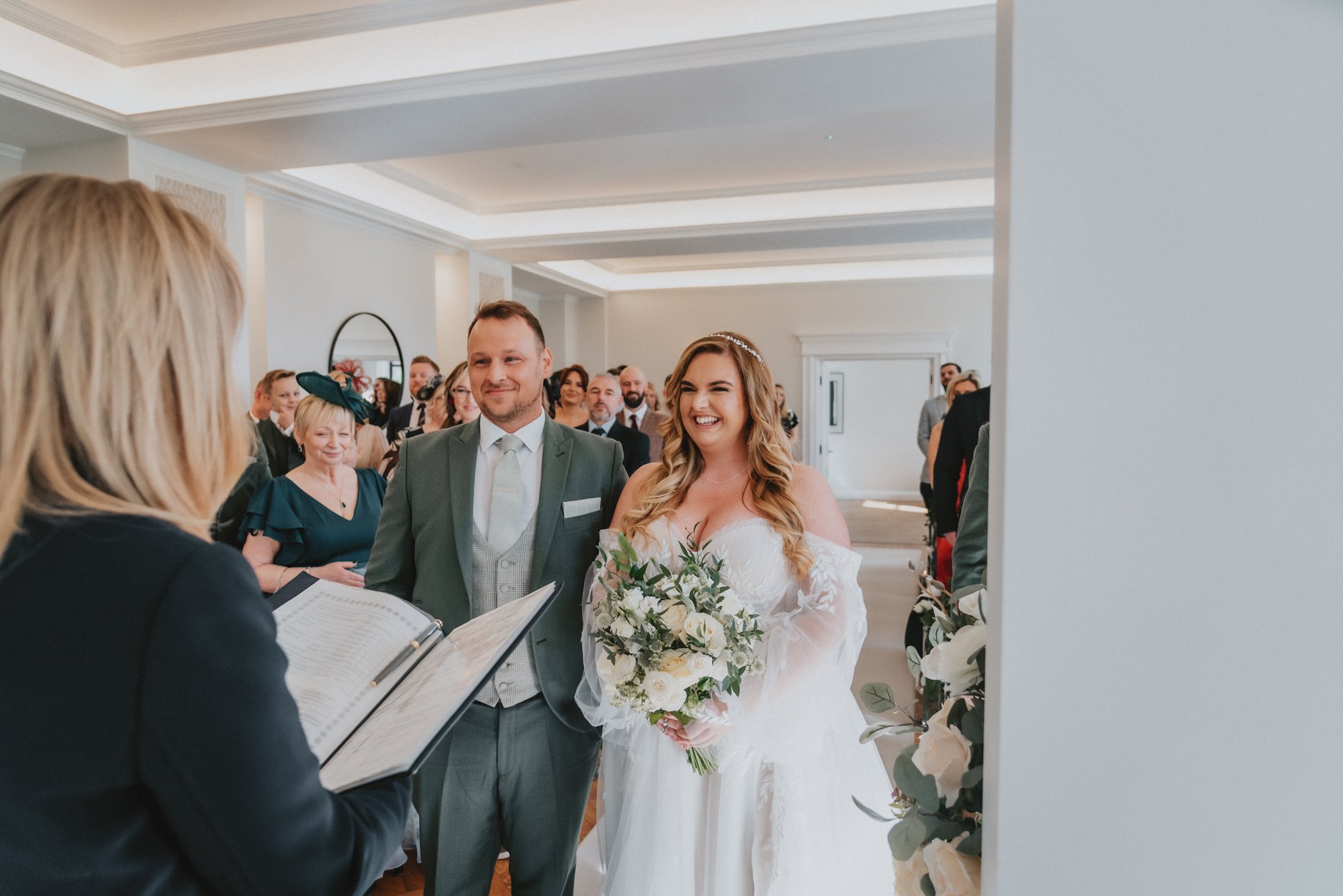 Bride and groom at their wedding ceremony, standing before an officiant, with guests in the background in an elegant indoor setting.