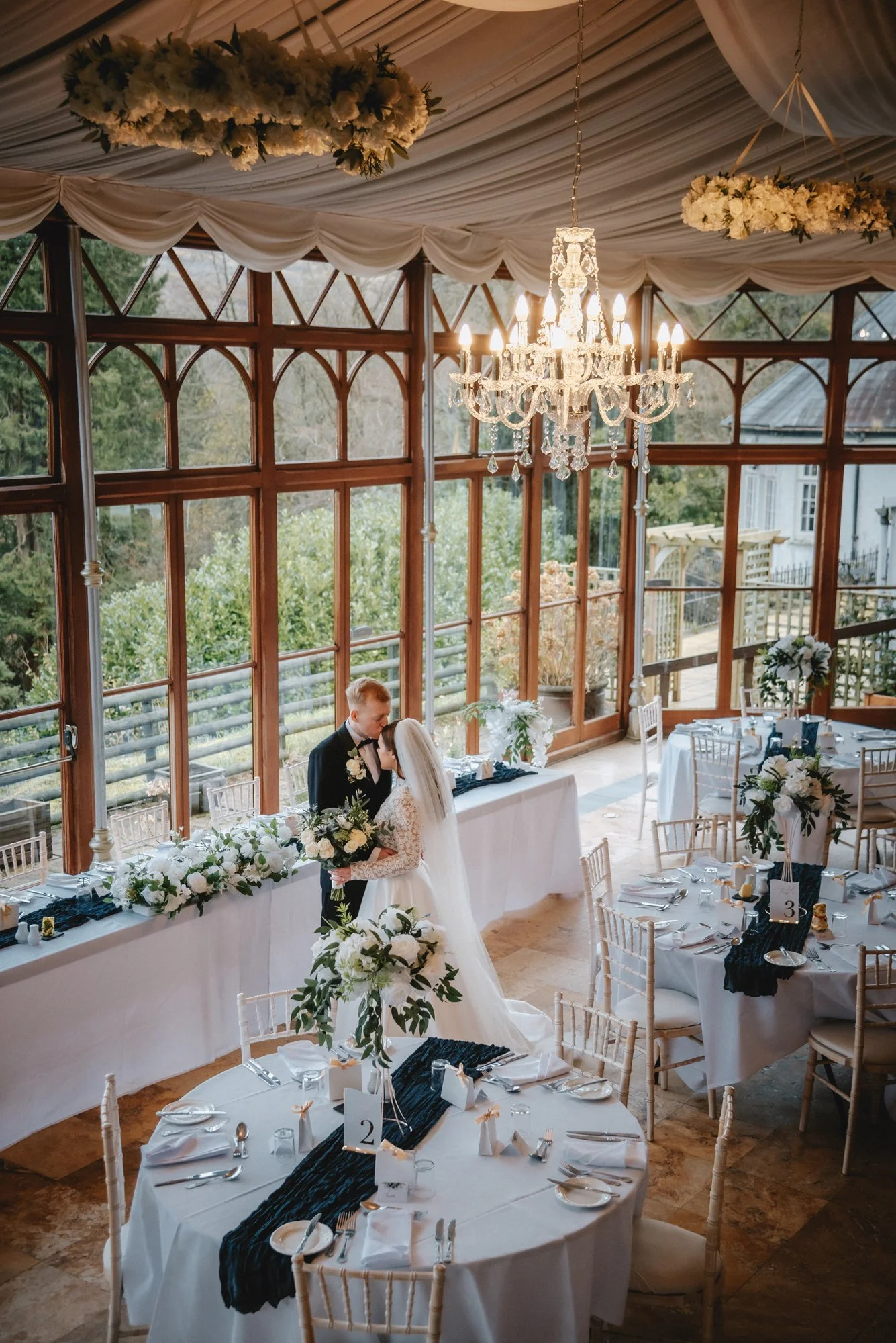 A bride and groom share a tender moment at their wedding reception, standing by a decorated table near large windows with a chandelier overhead, floral arrangements, and round tables set for guests.