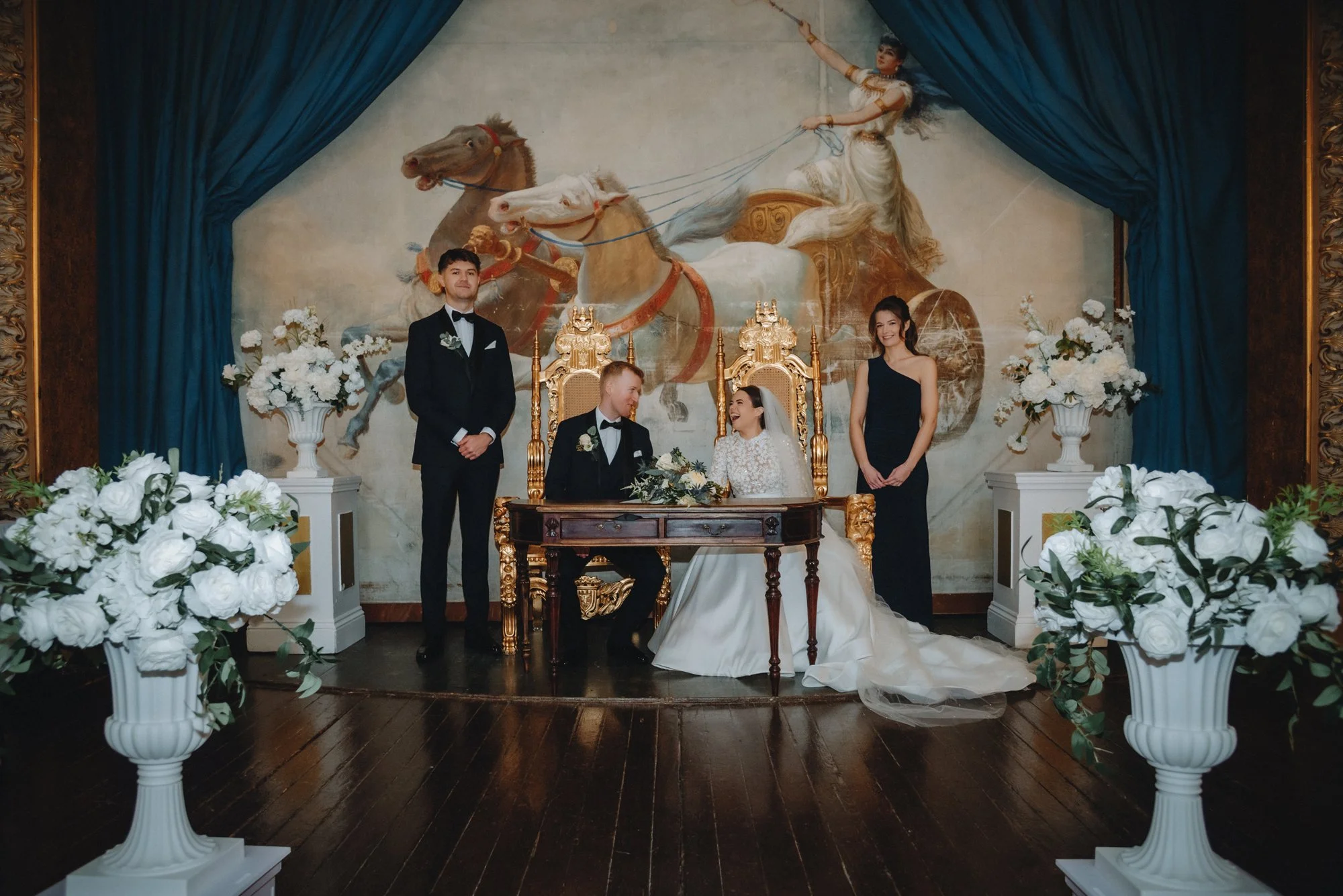 A wedding ceremony with a bride and groom sitting at a wooden table, surrounded by two groomsmen and a bridesmaid, in front of a large painting of a chariot with horses and a woman, with flower arrangements on white pedestals and blue curtains framin