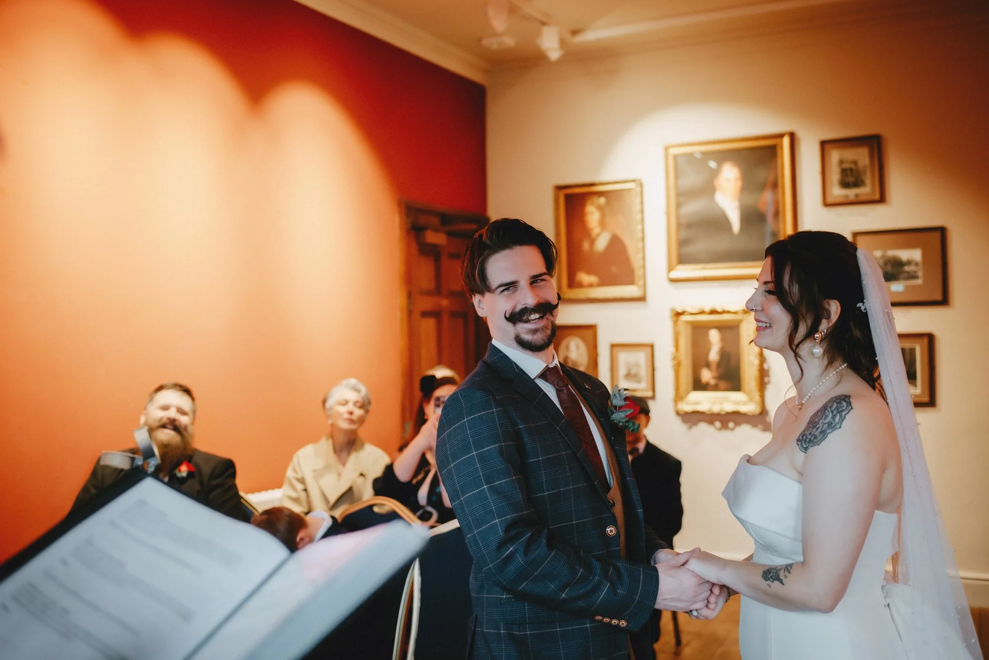 A bride and groom hold hands and smile during their wedding ceremony, with guests seated in the background in a warmly lit room with framed portraits on the wall.