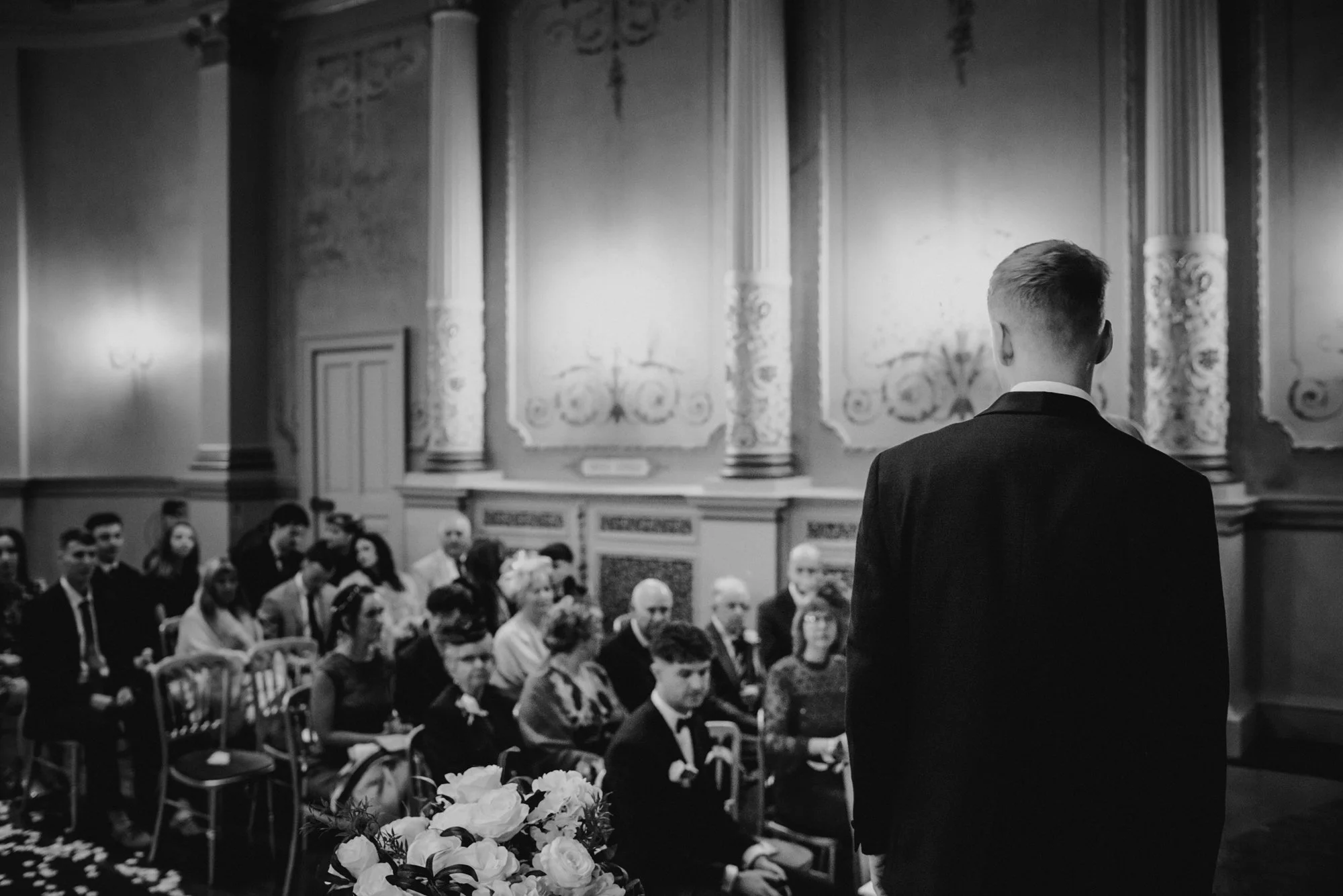 A man in a suit facing away, standing before a seated audience in an ornate room with decorative wall panels and floral arrangements.
