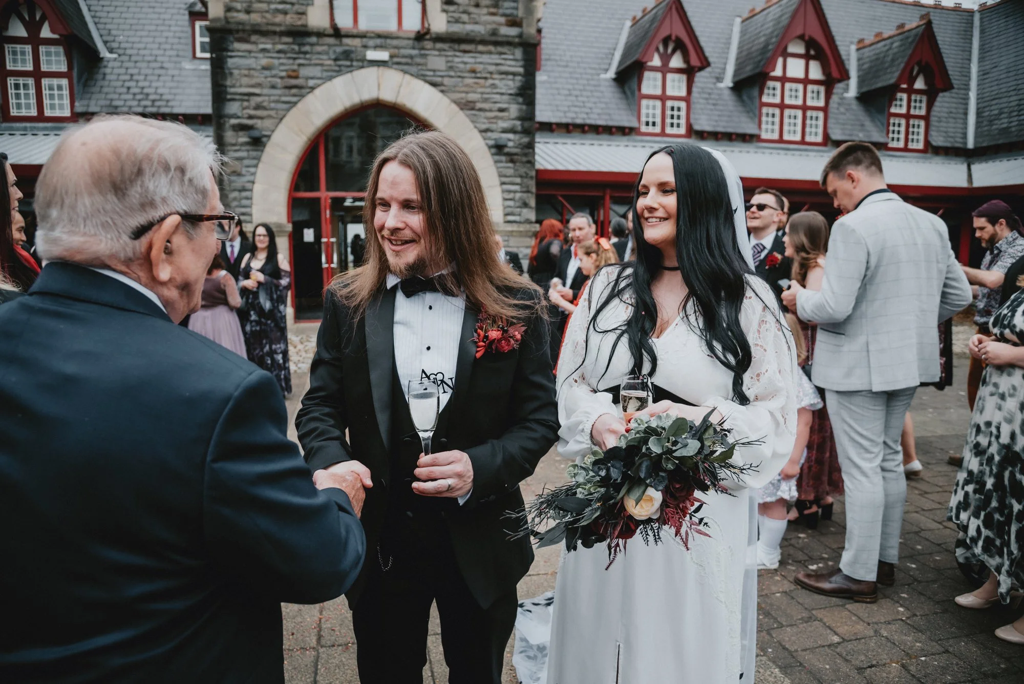 Bride and groom at their wedding reception, talking with an older man. The bride holds a bouquet and both are smiling. The groom wears a tuxedo, and the scene is outdoors with other guests in the background.