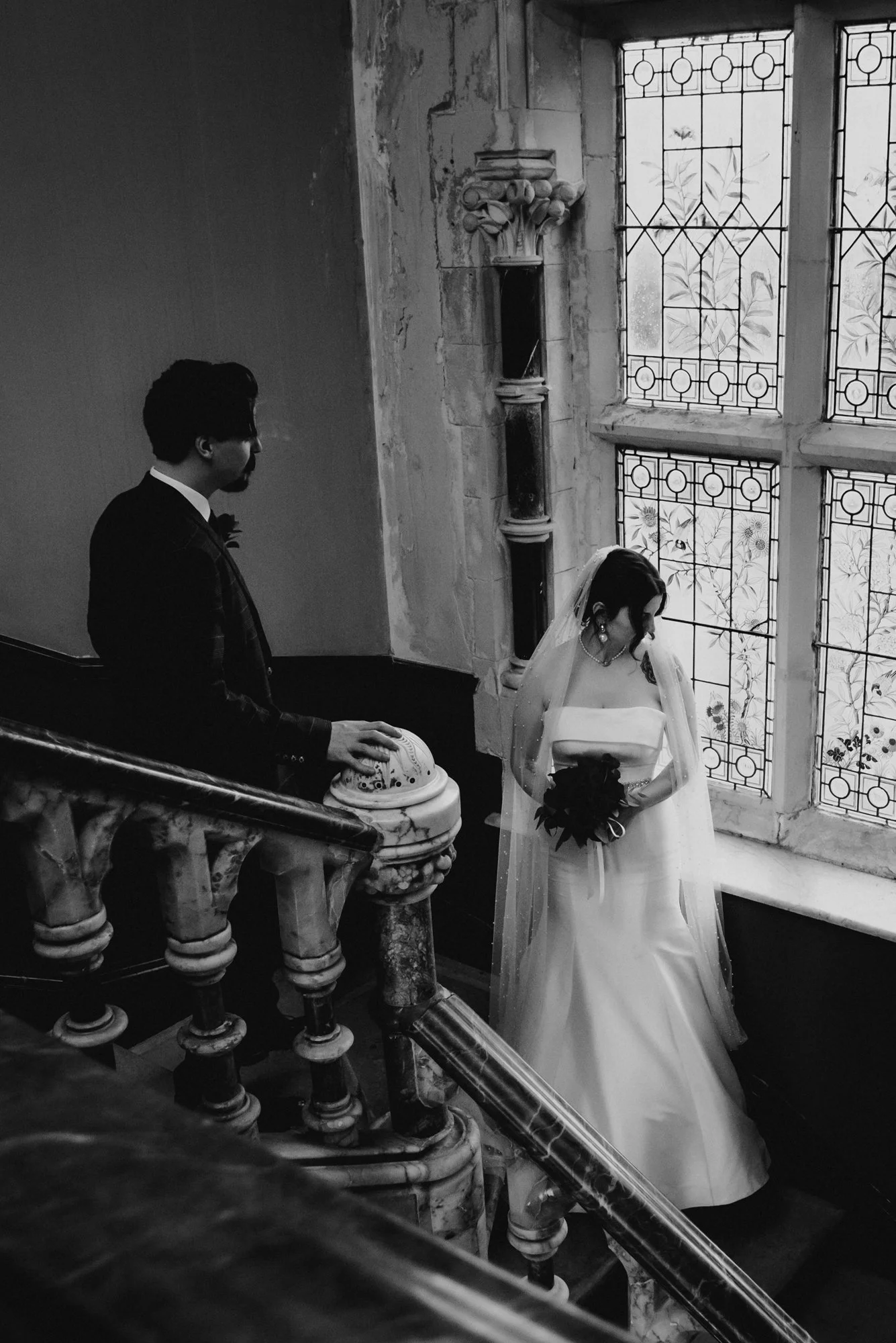 A black and white photo of a bride in a wedding dress holding a bouquet looking down, standing near a large window with decorative stained glass, with a groom in a suit standing near a carved marble staircase railing, inside a historic church or cath