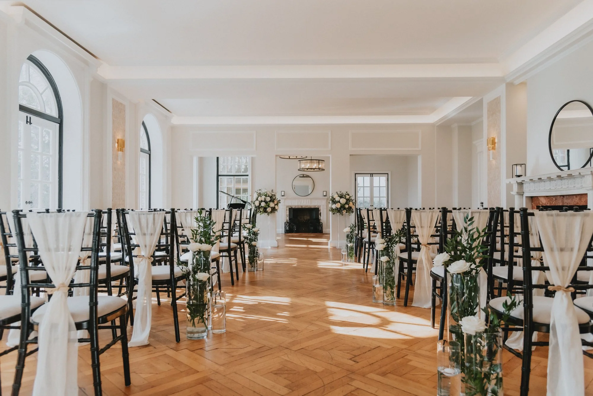 Elegant wedding ceremony setup in a bright, spacious room with black chairs decorated with white fabric and flowers, and a fireplace at the front.