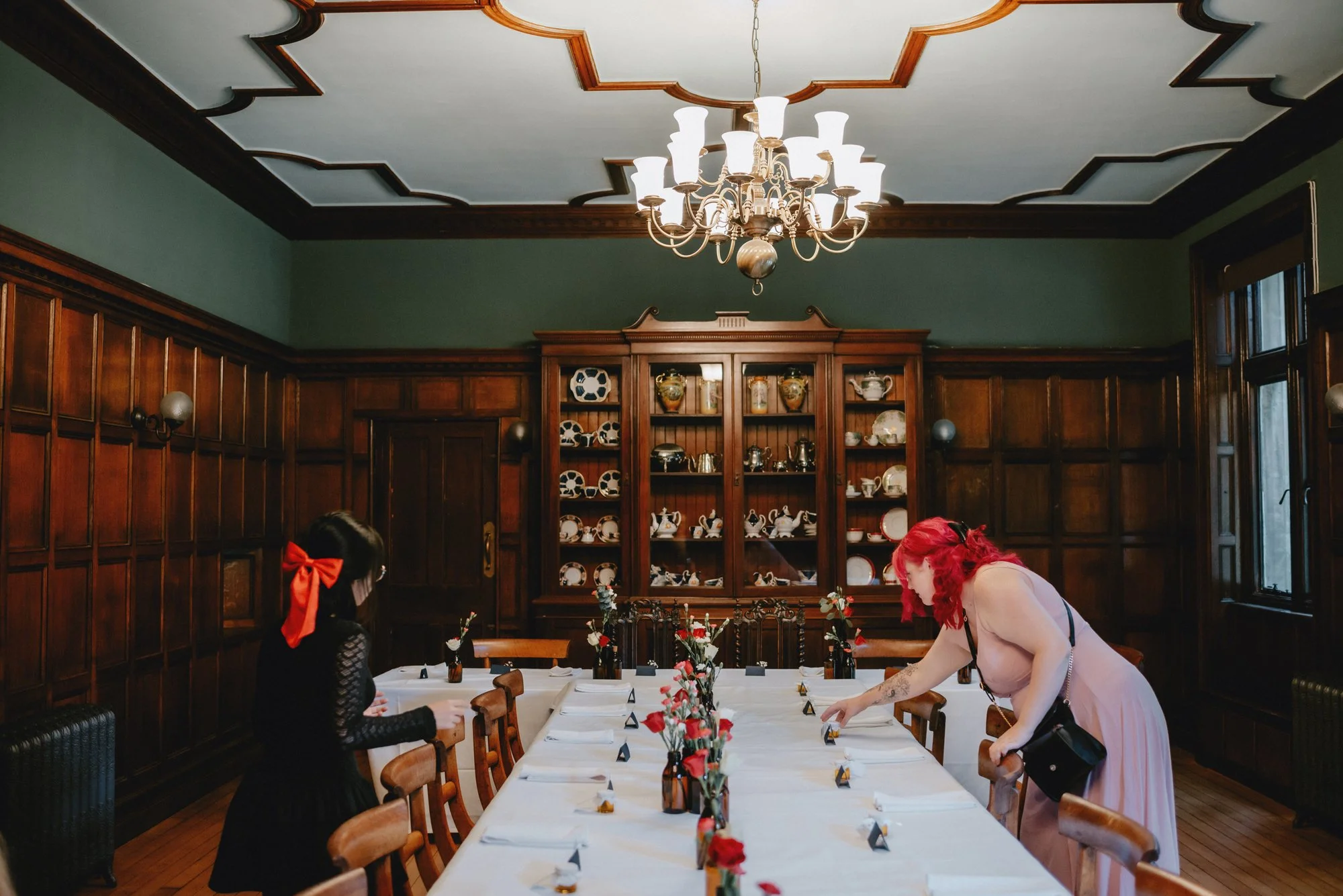 A dining room with dark wood paneled walls, a large chandelier, and a long table set for a meal with white tablecloths and floral centerpieces. Two women are in the room, one with red hair in a pink dress and the other with dark hair and a red bow, b