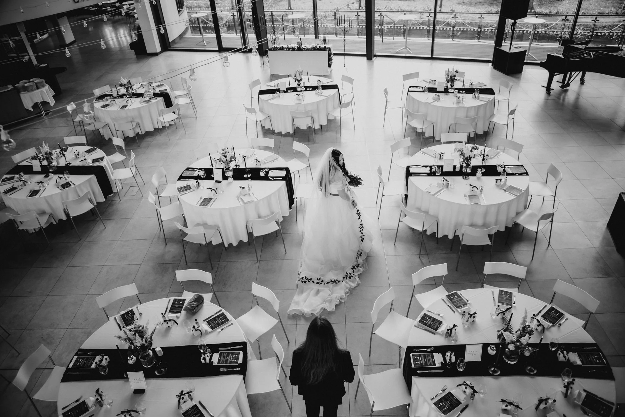 A wedding reception setup with round tables decorated with floral centerpieces, place settings, and chairs arranged around them. There is a bride in a wedding gown and a person standing nearby in the center of the hall. Large windows in the backgroun