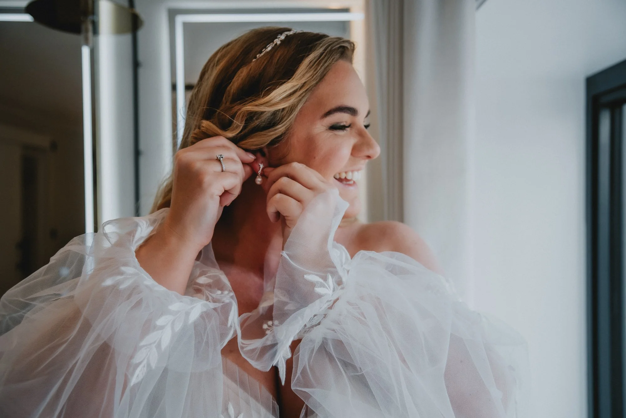 A woman, likely a bride, smiling and putting on an earring while looking out a window. She is wearing a white, tulle, puffy sleeve dress and has blonde hair styled with a headband.