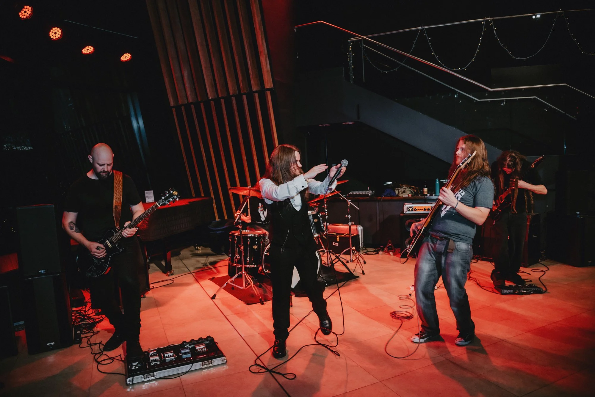 A band performs on stage with a female vocalist, guitarist, bassist, drummer, and another guitarist in a dimly lit venue with red and orange lighting.