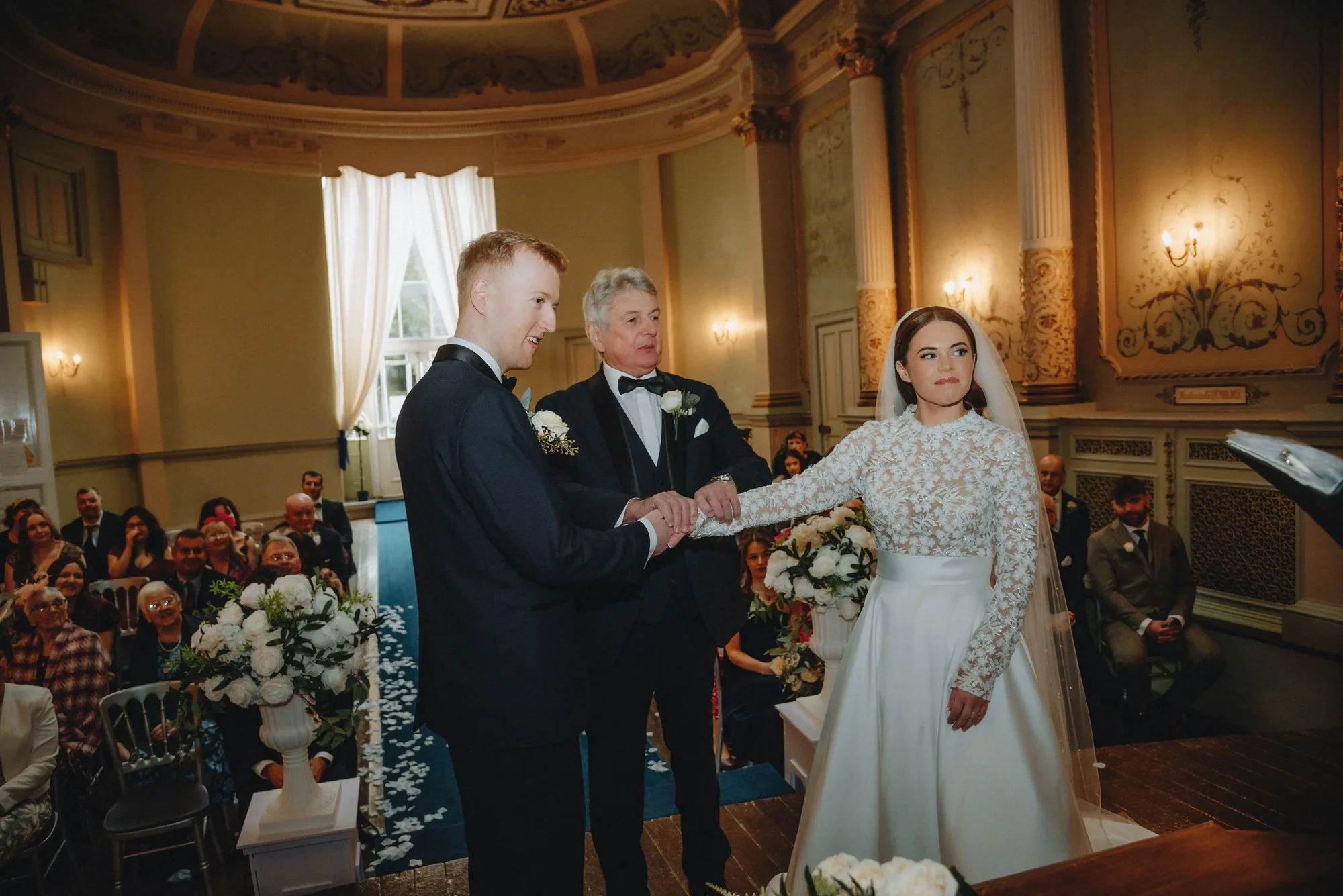 A wedding ceremony taking place indoors with a bride, groom, and officiant holding hands. Guests seated in the background, floral arrangements, and elegant decor visible.