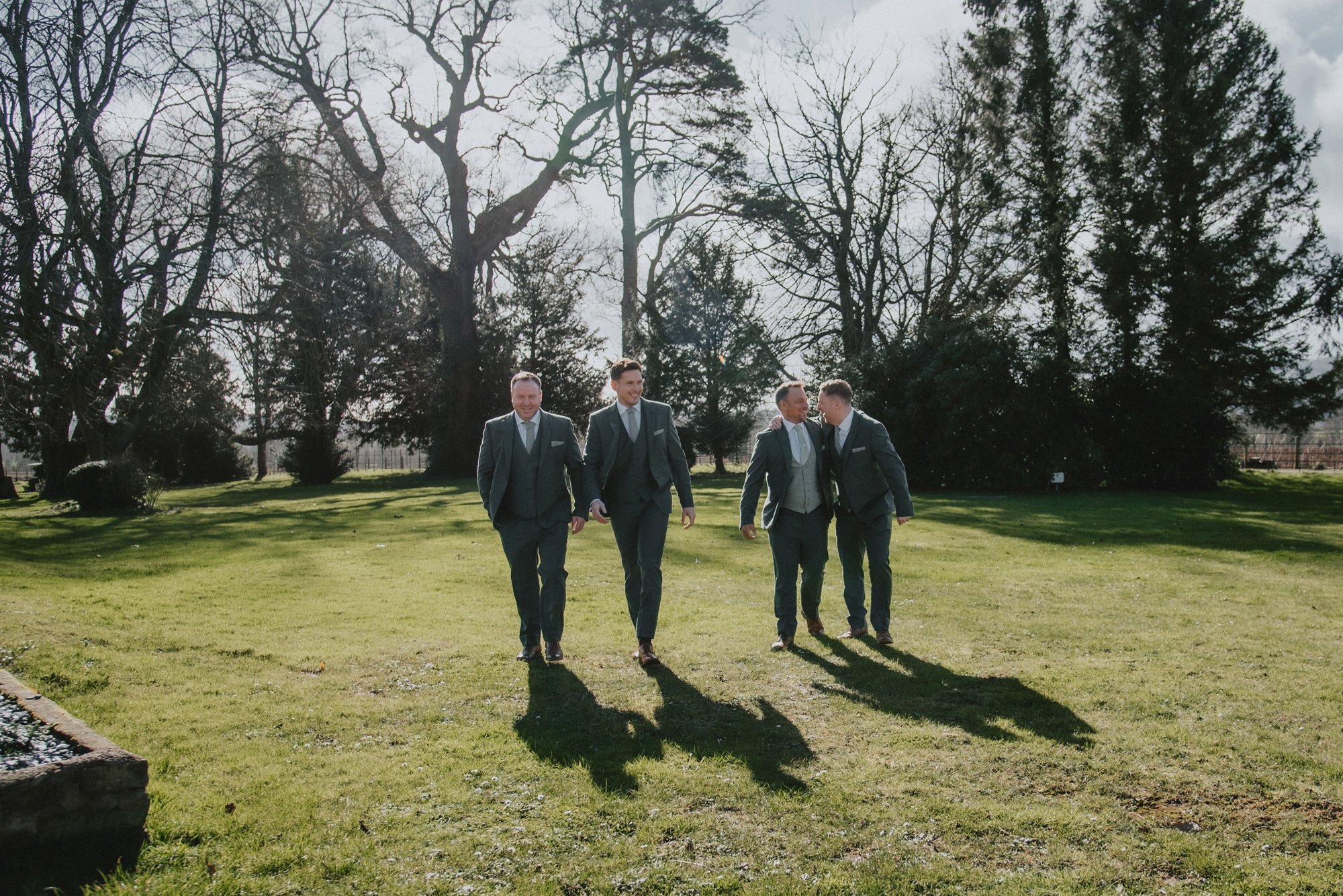 Four men in suits walking and talking outdoors on a sunny day, with leafless trees in the background and shadows cast on the grass.