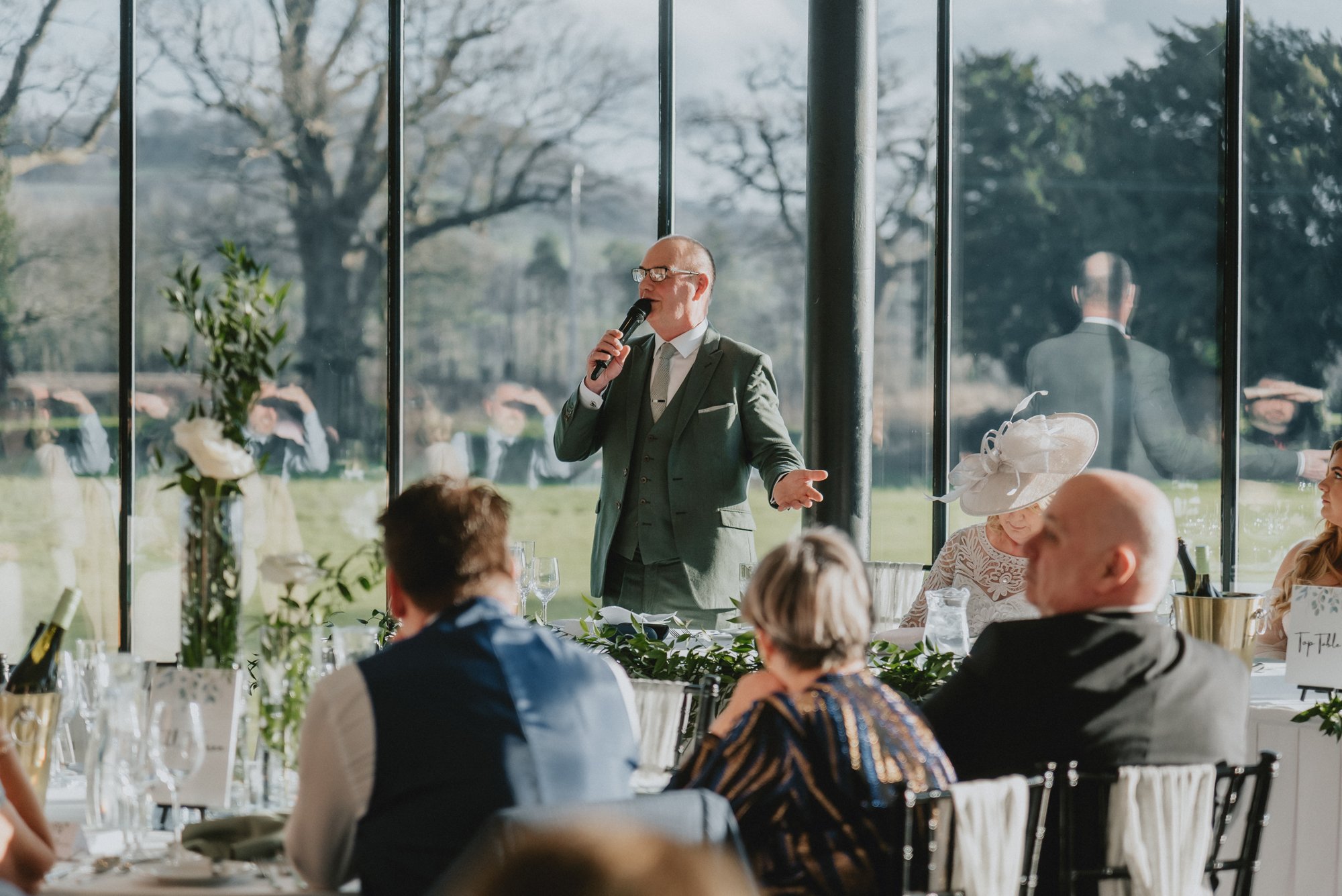 A man in a green suit giving a speech at a wedding reception, standing in front of large windows with a scenic outdoor view, while guests sit at tables and listen.
