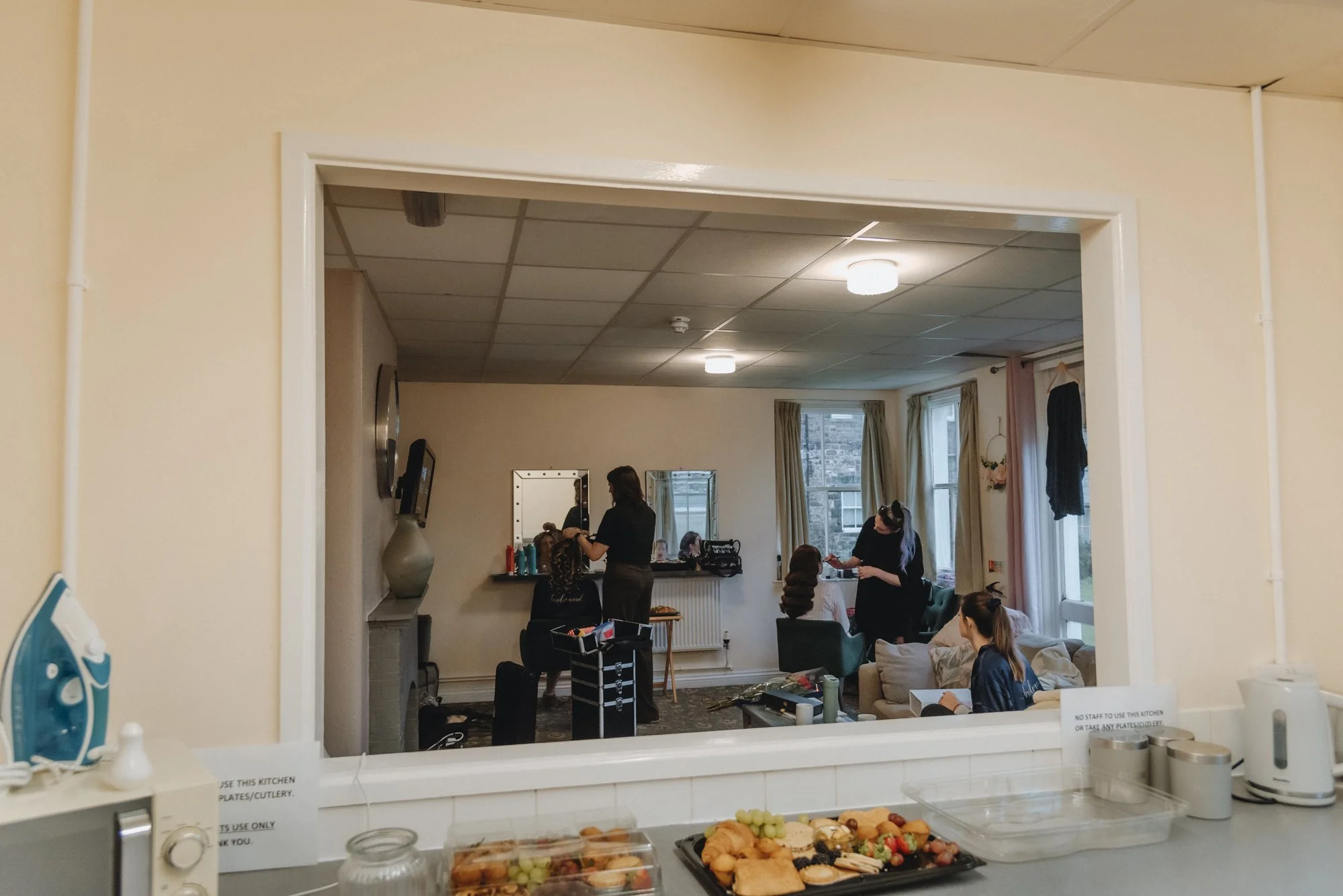 View of a busy room seen through a kitchen window. The room is decorated with beige walls and curtains, with three women styling hair, and a woman working on a laptop, all seated or standing near mirrors. The space has large windows letting in natura